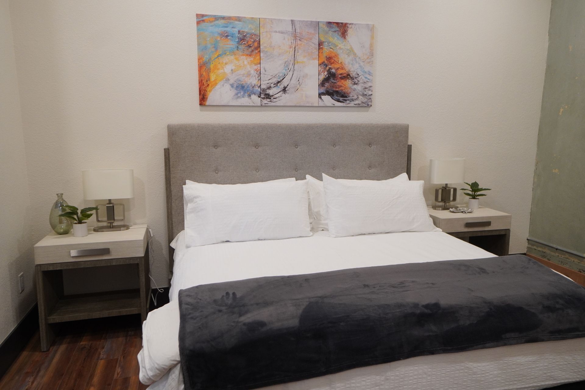 Bedroom with bed, nightstands, artwork, and plants. White bedding, gray headboard and blanket. Wooden floor.