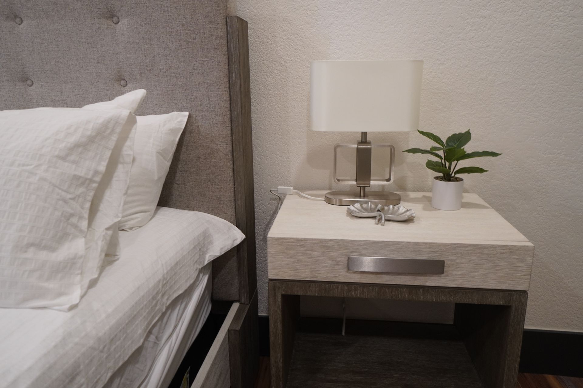 Bed with white linens next to a nightstand with a lamp and small plant.