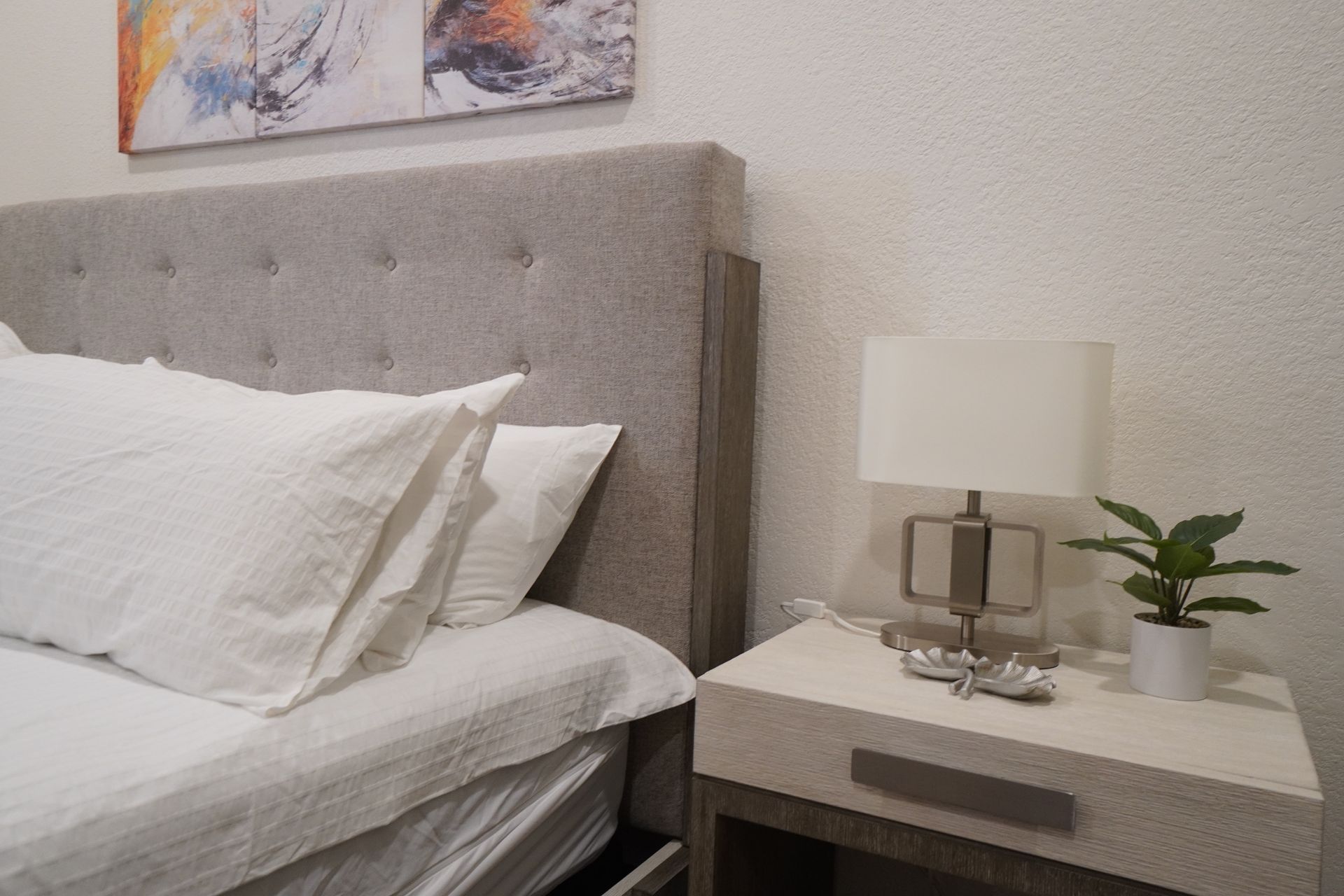 Bedroom with a bed, nightstand with lamp and plant. Artwork hangs above bed.