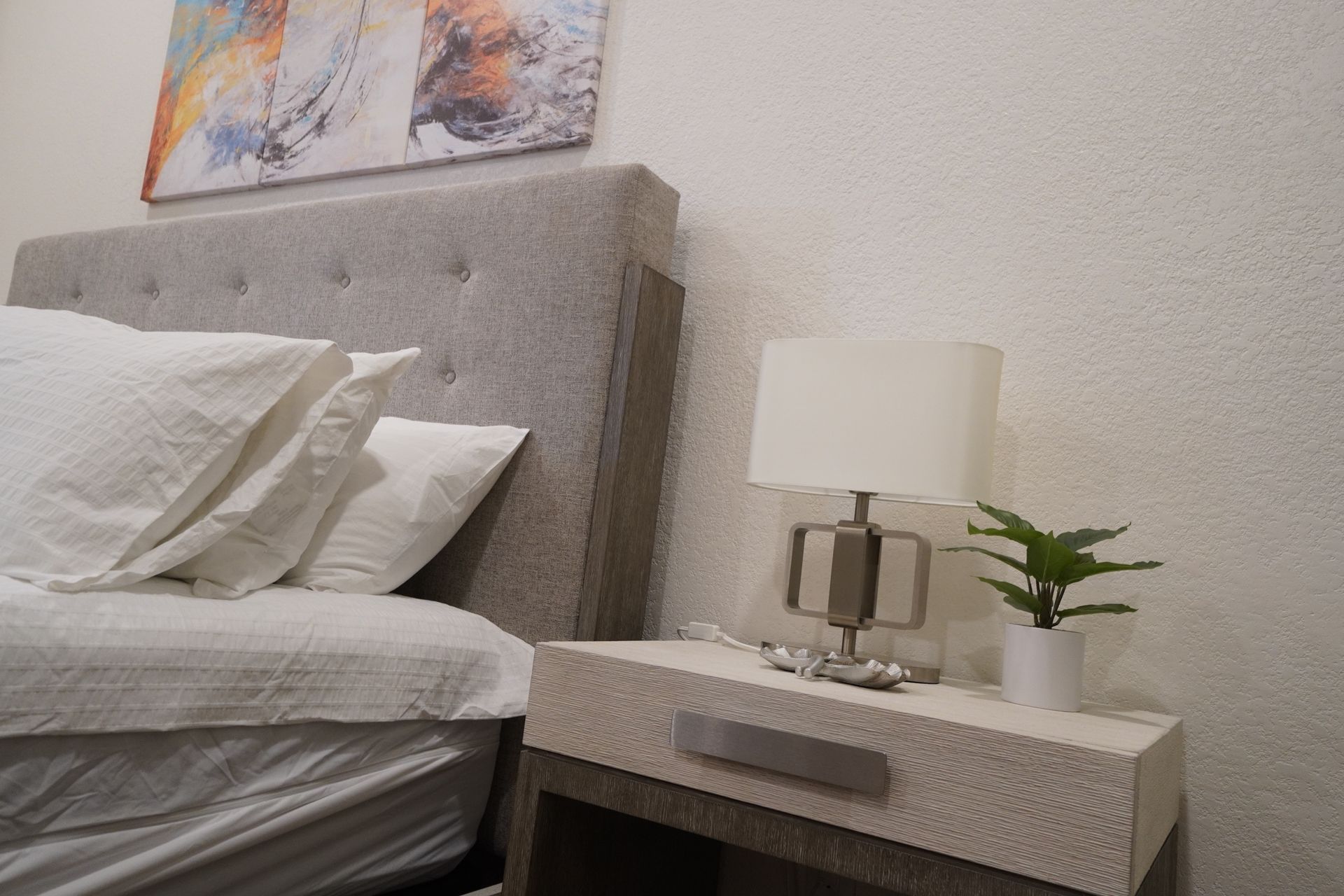 Bedroom scene with gray upholstered headboard, white bedding, and a nightstand with a lamp and small plant.