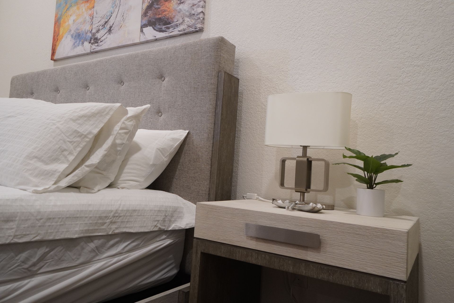Bedroom scene: Gray tufted headboard, white bedding, lamp on bedside table, small plant.