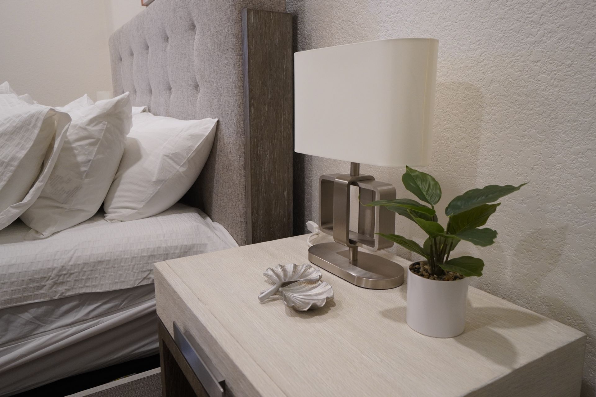 Bedroom scene: bed with pillows, nightstand with lamp, plant, and decorative dish.