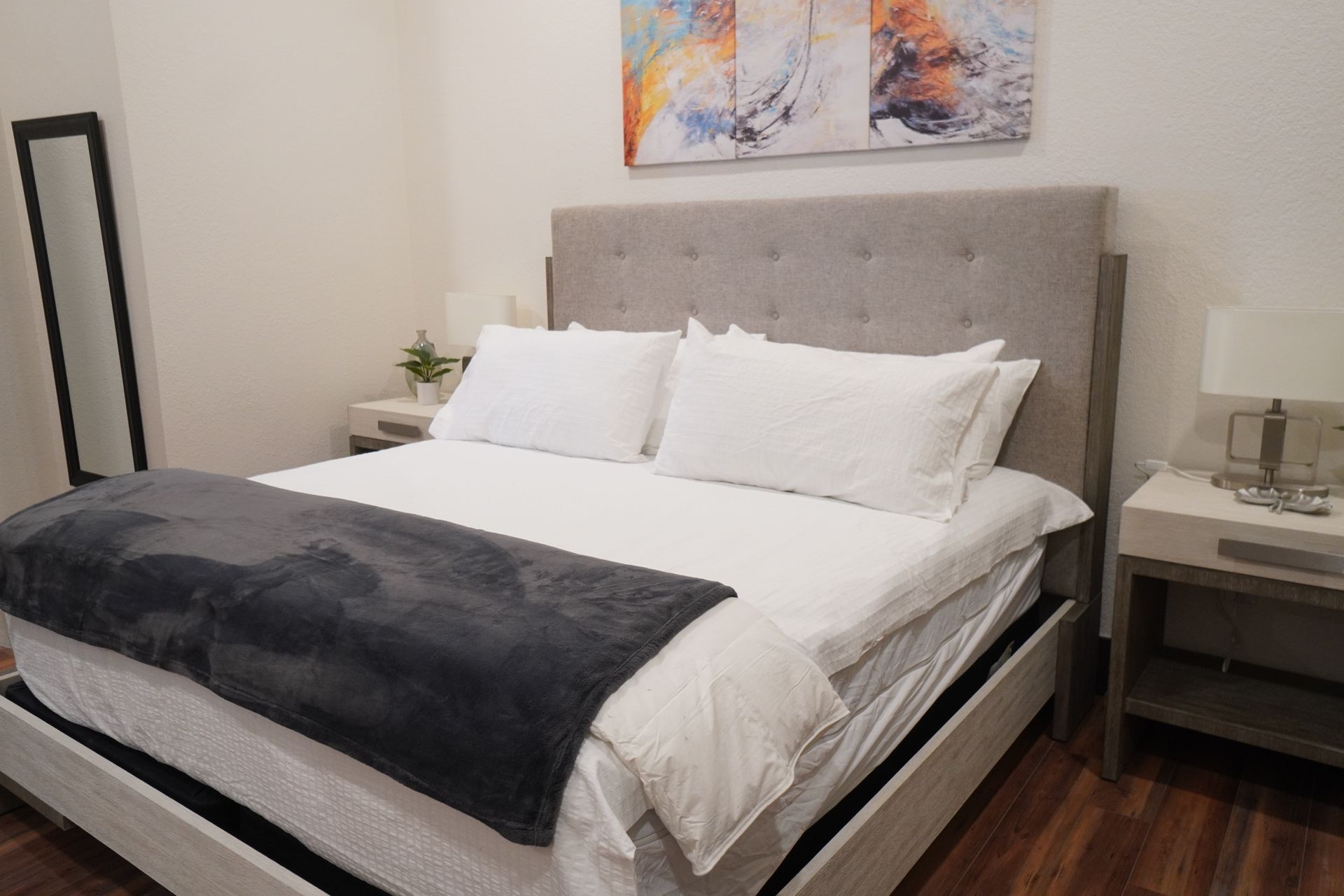 Bedroom with a large bed, gray headboard, white bedding, and a gray blanket. Includes a full-length mirror and a painting.