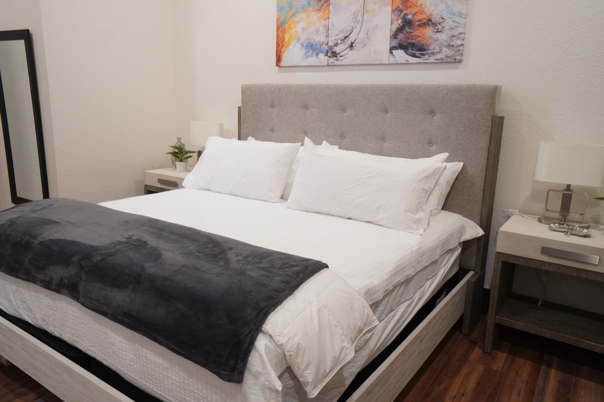 Bedroom with a bed, nightstands, and artwork on the wall. The bed has white bedding and a gray throw.