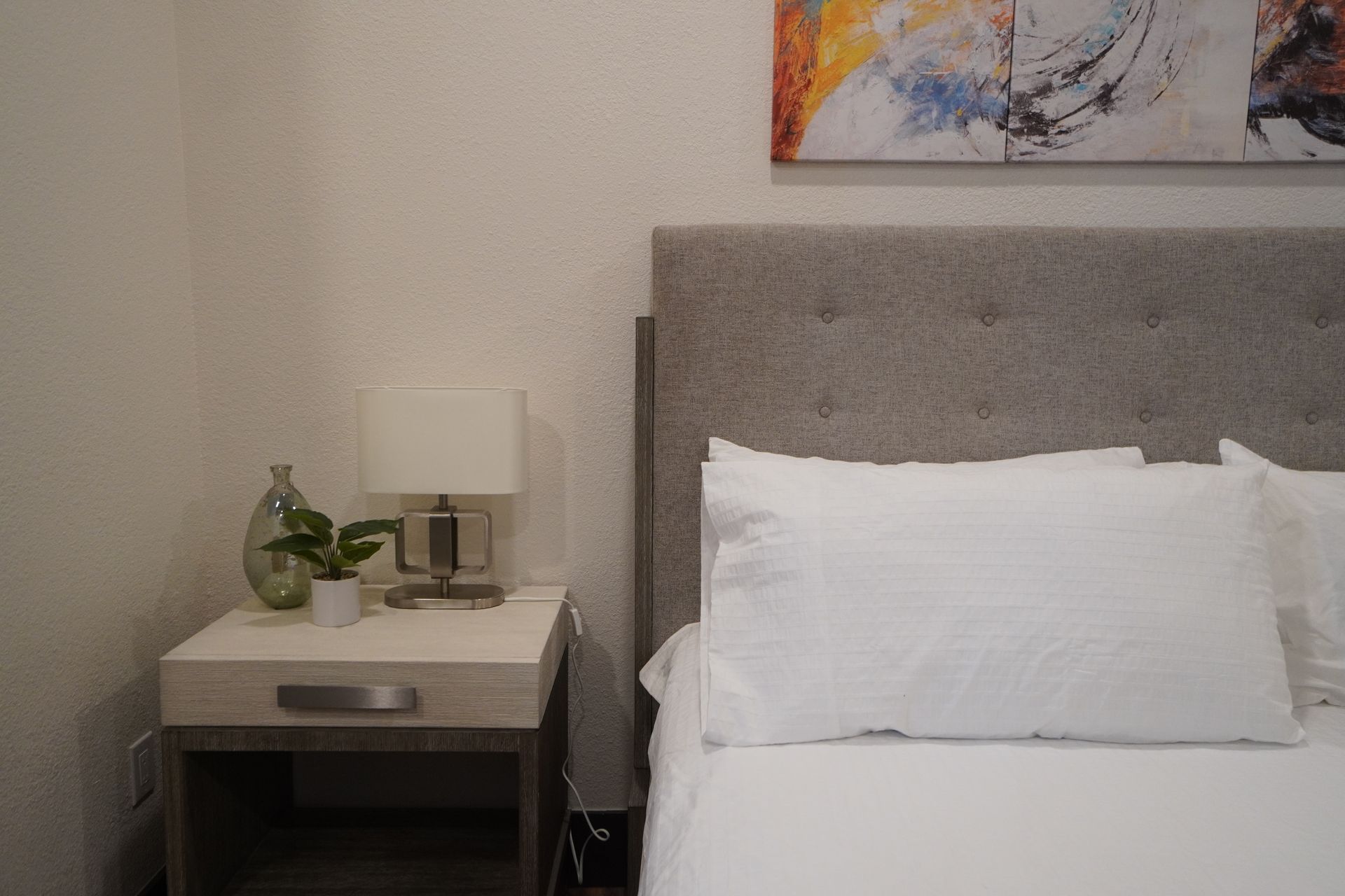 Bedside table with lamp, art, and white bedding.