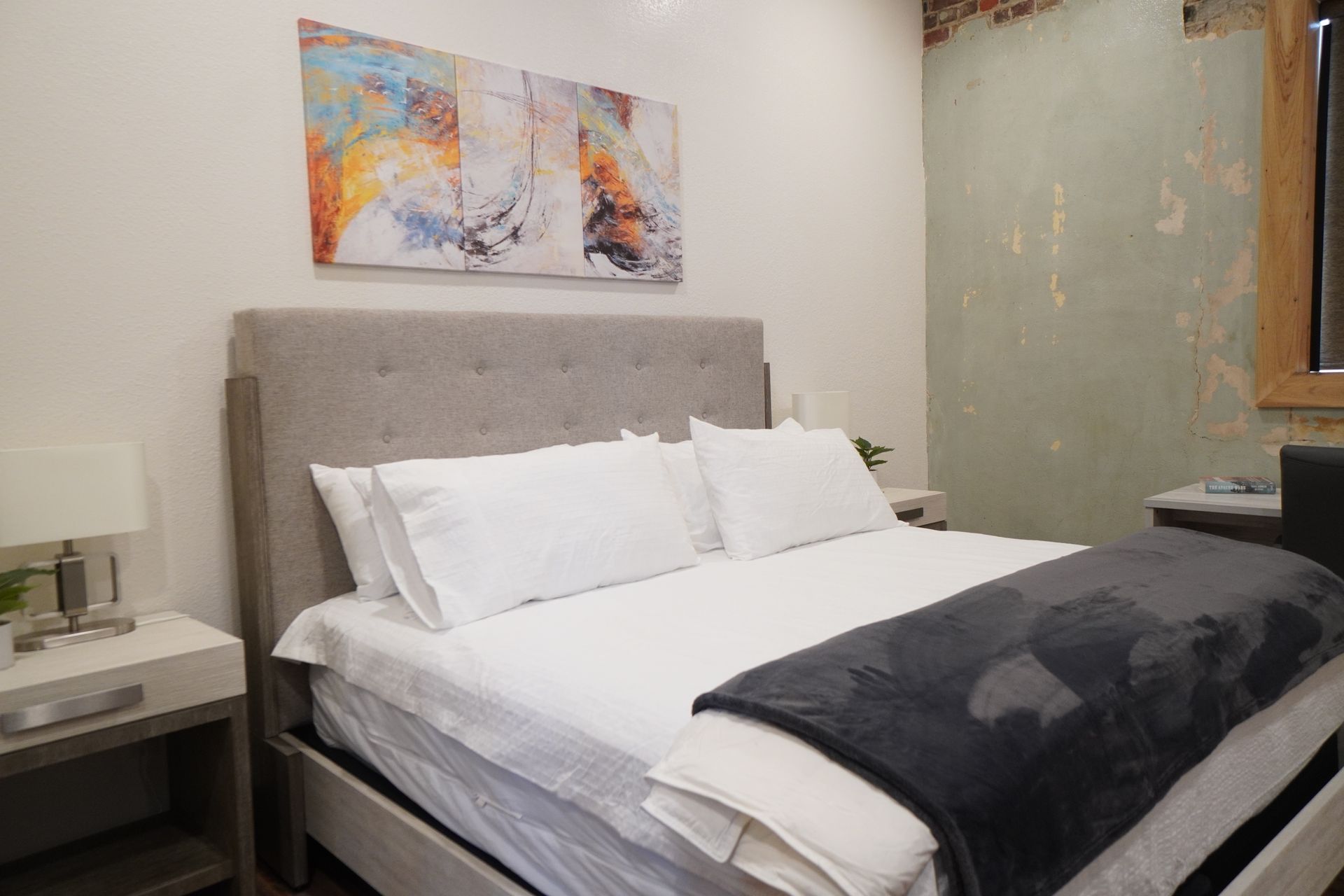 Bedroom with a large bed, abstract artwork, bedside table, and a wall with exposed brick.