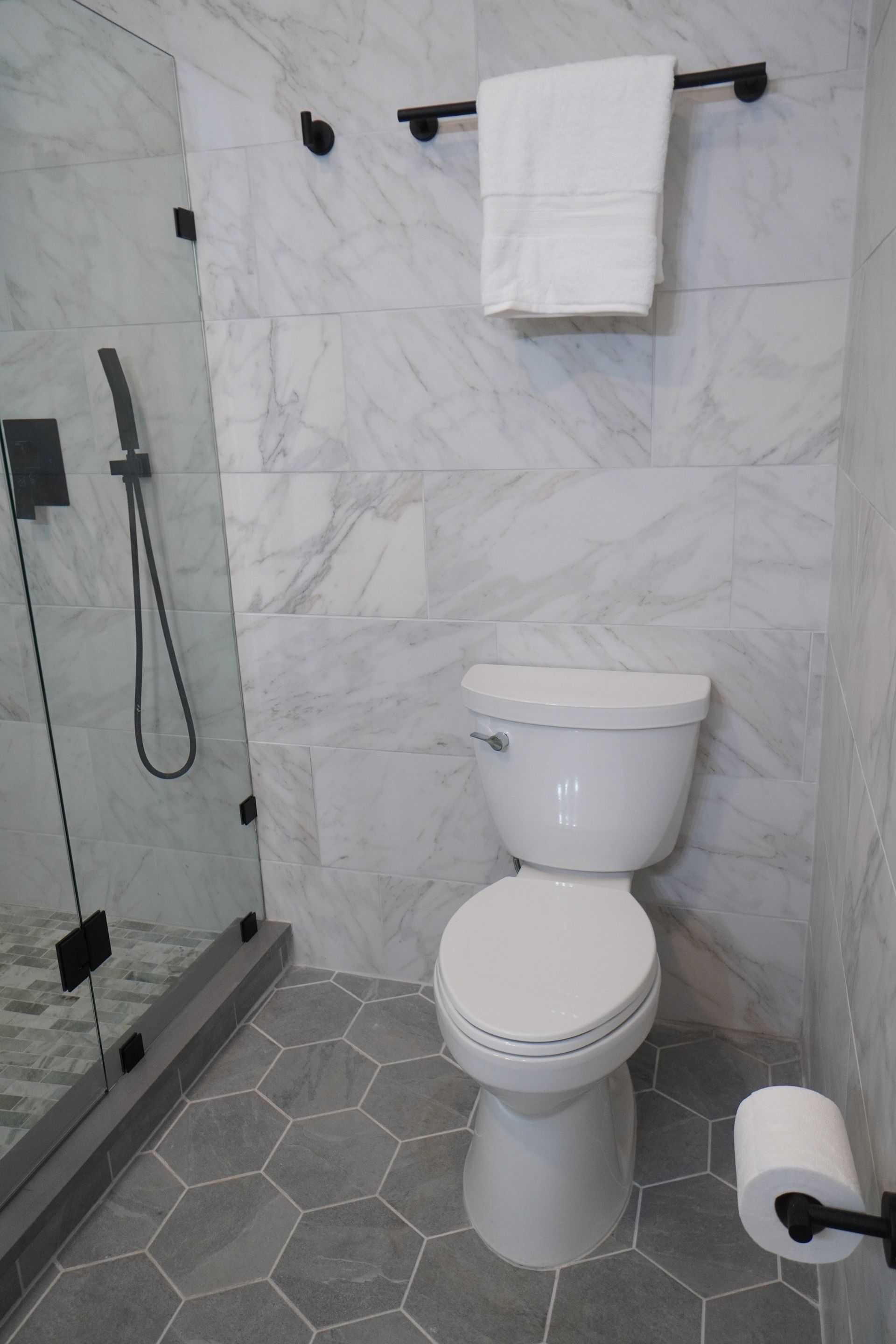 Modern bathroom with white toilet, marble tile, gray hexagon floor, and black fixtures.