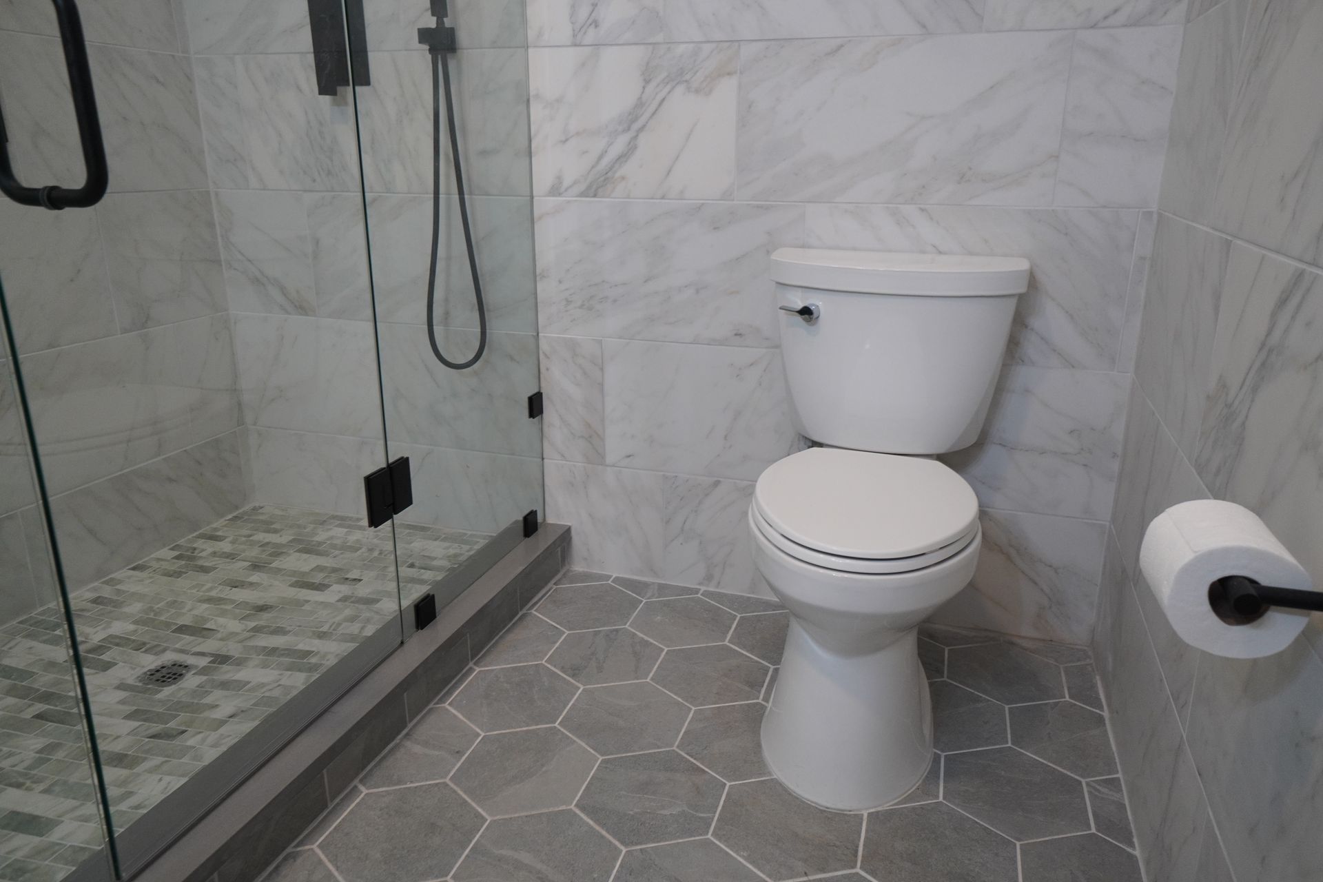 Bathroom with gray hexagon floor tiles, marble walls, and a white toilet. A glass shower is on the left.