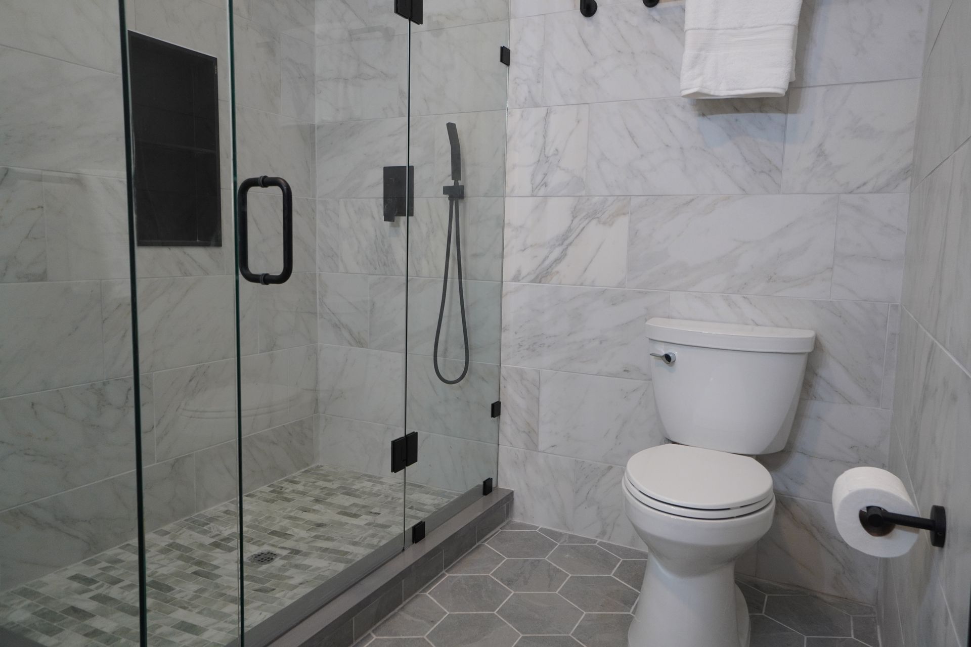 Bathroom with glass shower, white toilet, gray hexagonal floor tiles, and marble-like wall tiles.