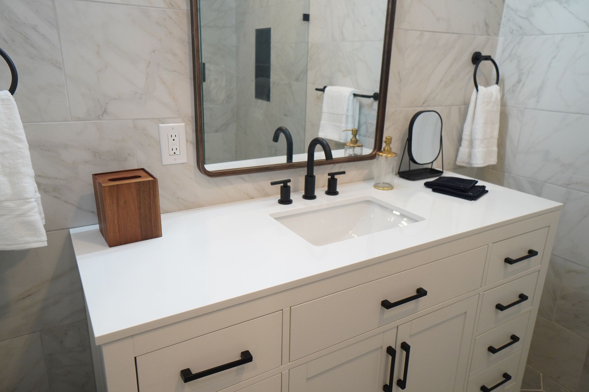 Bathroom vanity with white countertop, black fixtures, and mirror. White towels hang on black towel rings.