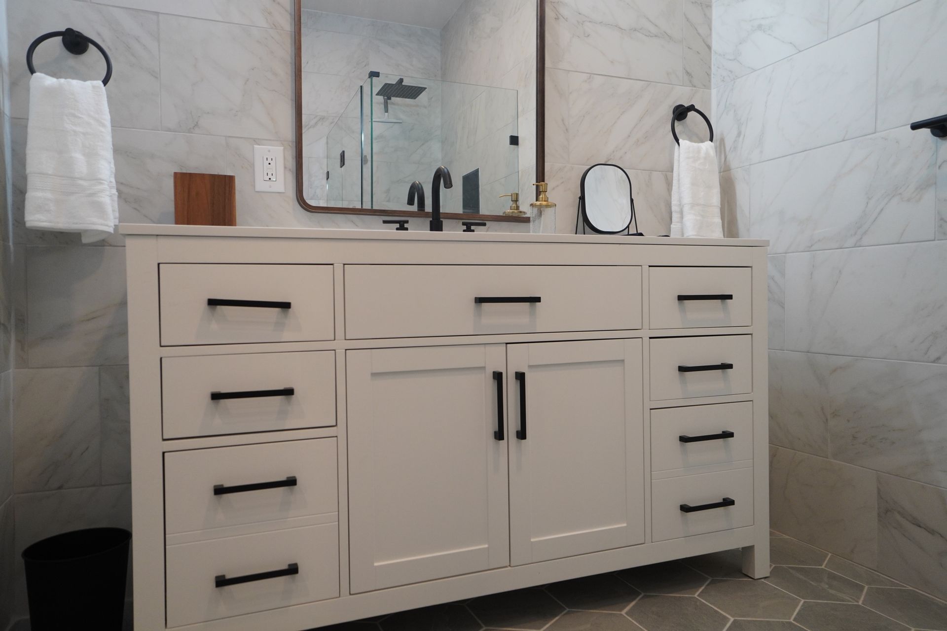 Bathroom vanity with white cabinet, black hardware, mirror, and towel rings; gray tile walls and floor.