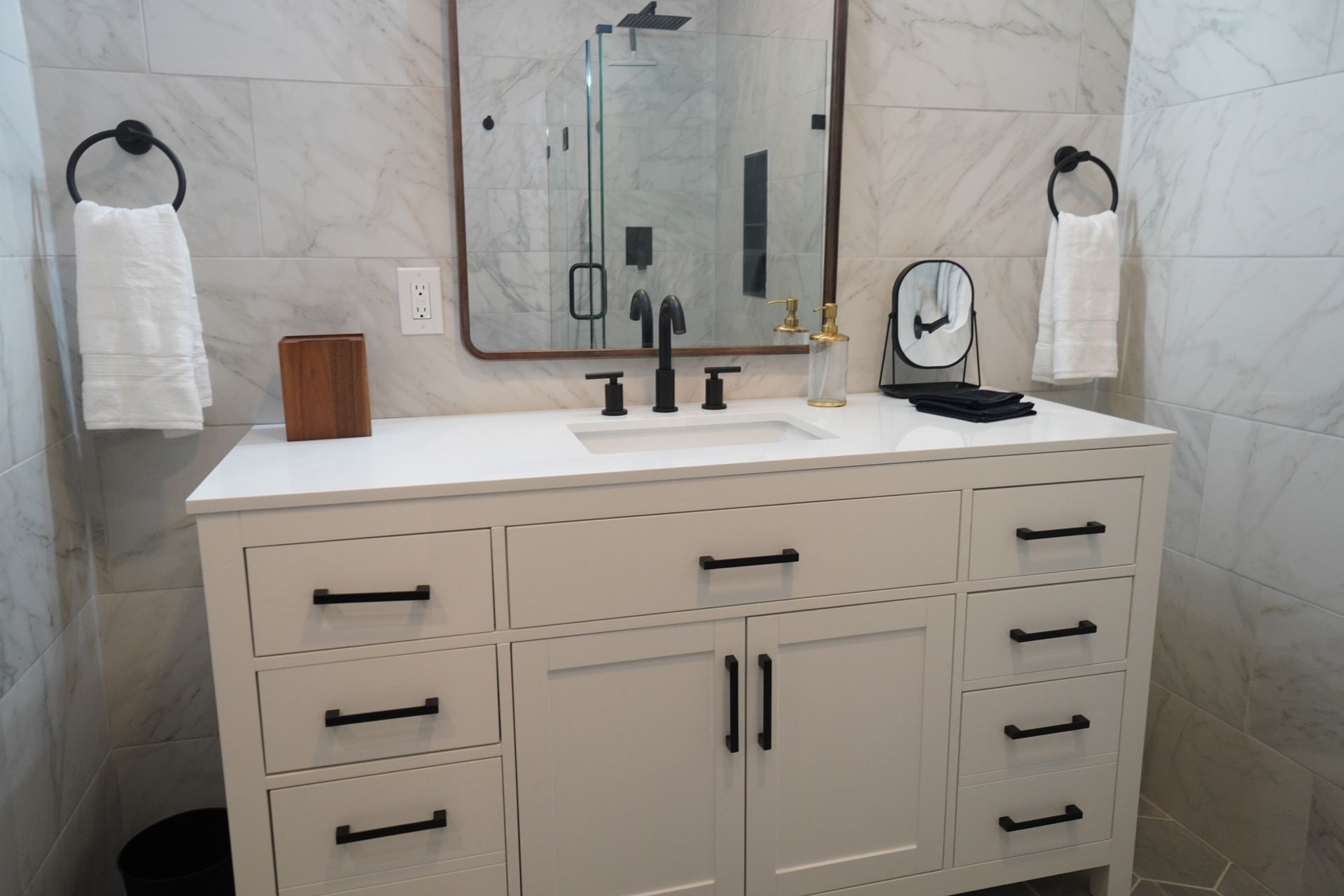 White bathroom vanity with black hardware, mirror, towels, and marble tile.