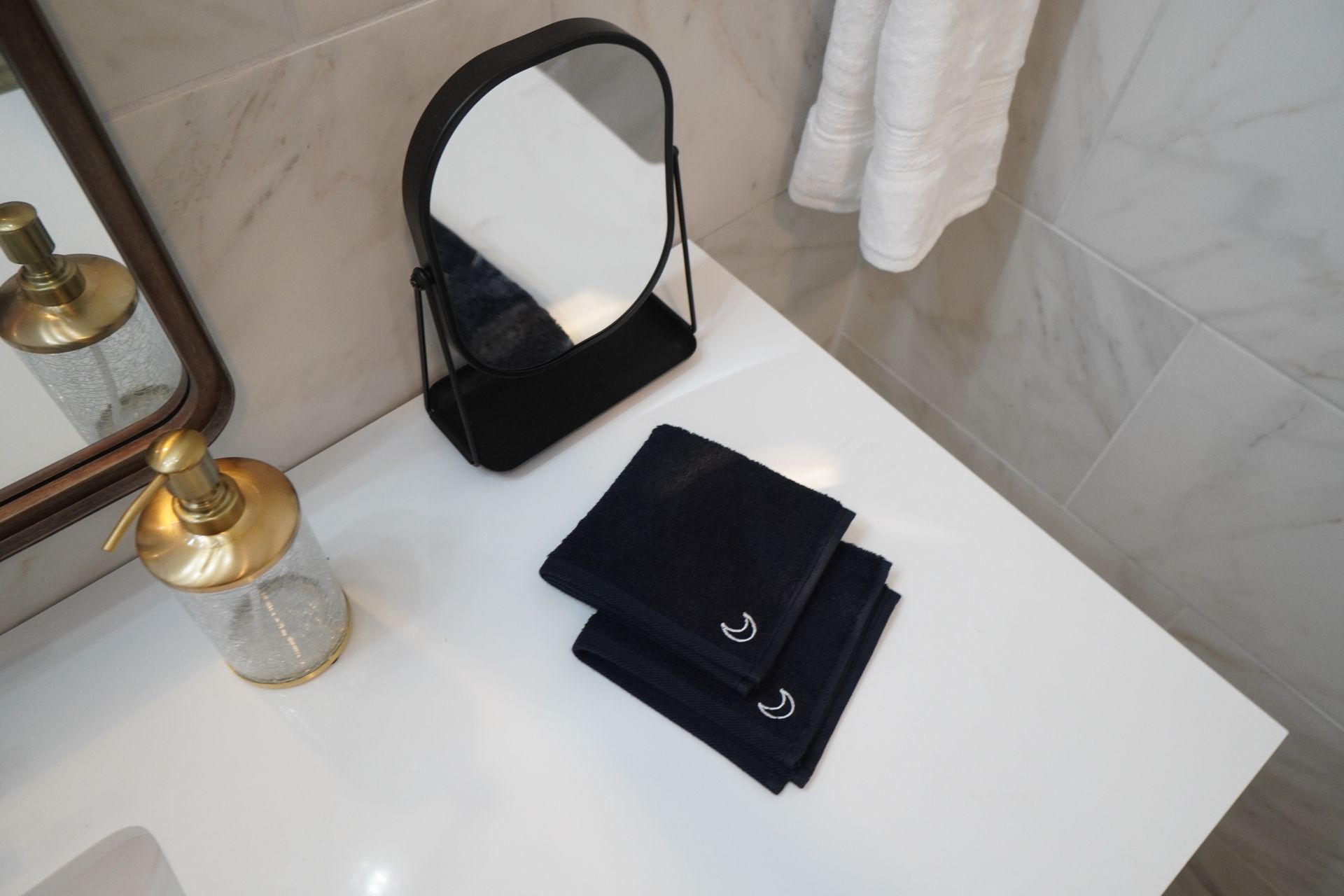 Black towels, mirror, and soap dispenser on a white countertop in a bathroom.