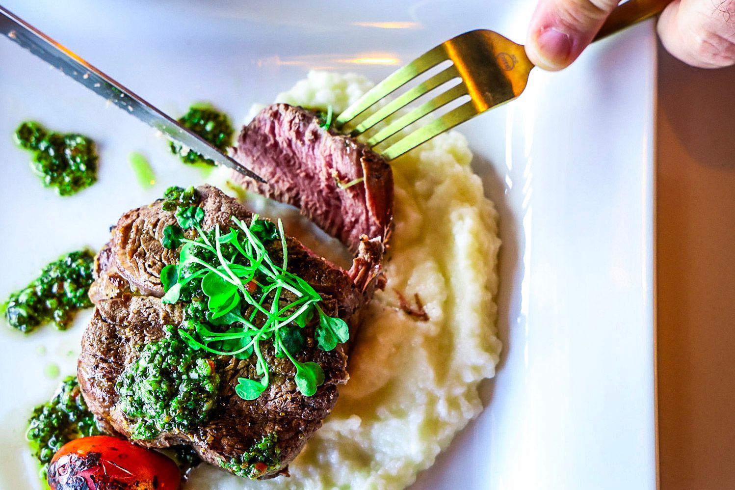 Steak on mashed potatoes with green sauce, cut by a knife and fork on a white plate.