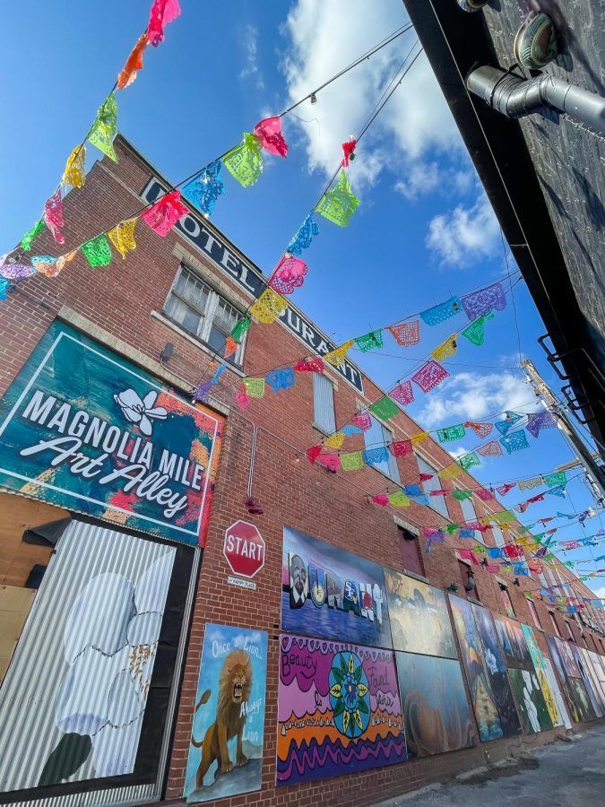 Magnolia Mile Art Alley with colorful murals, flags, and a brick building under a partly cloudy sky.