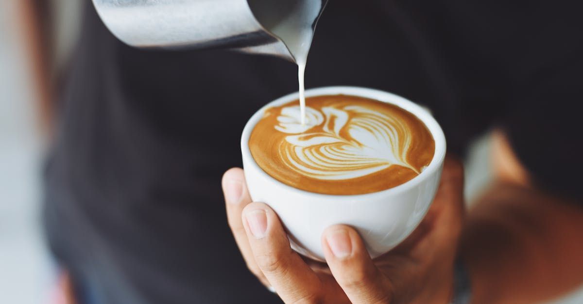 Barista pouring milk into coffee, creating a latte art design of a leaf in a white mug.