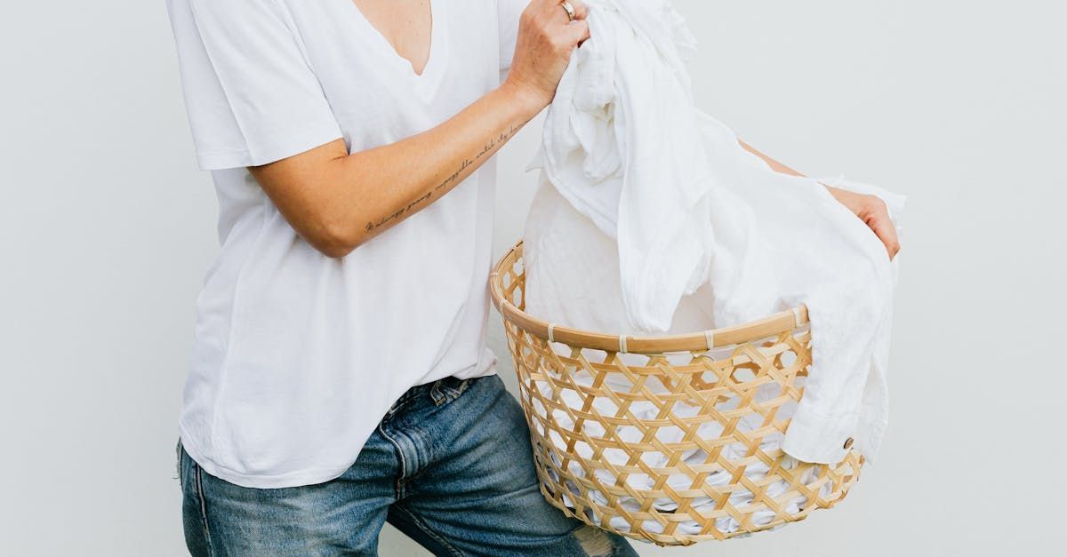 Person in white shirt and jeans holding white laundry in a wicker basket against a white wall.
