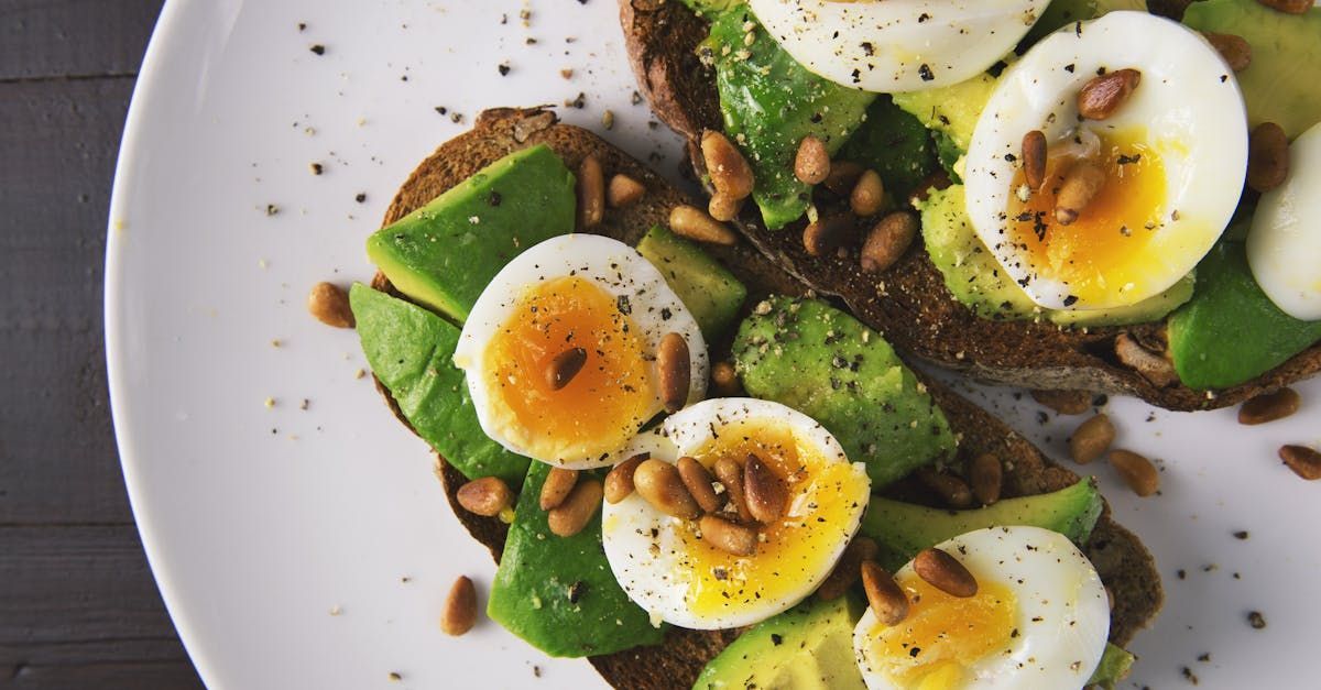 Avocado toast with sliced boiled eggs and pine nuts, sprinkled with black pepper, on a white plate.