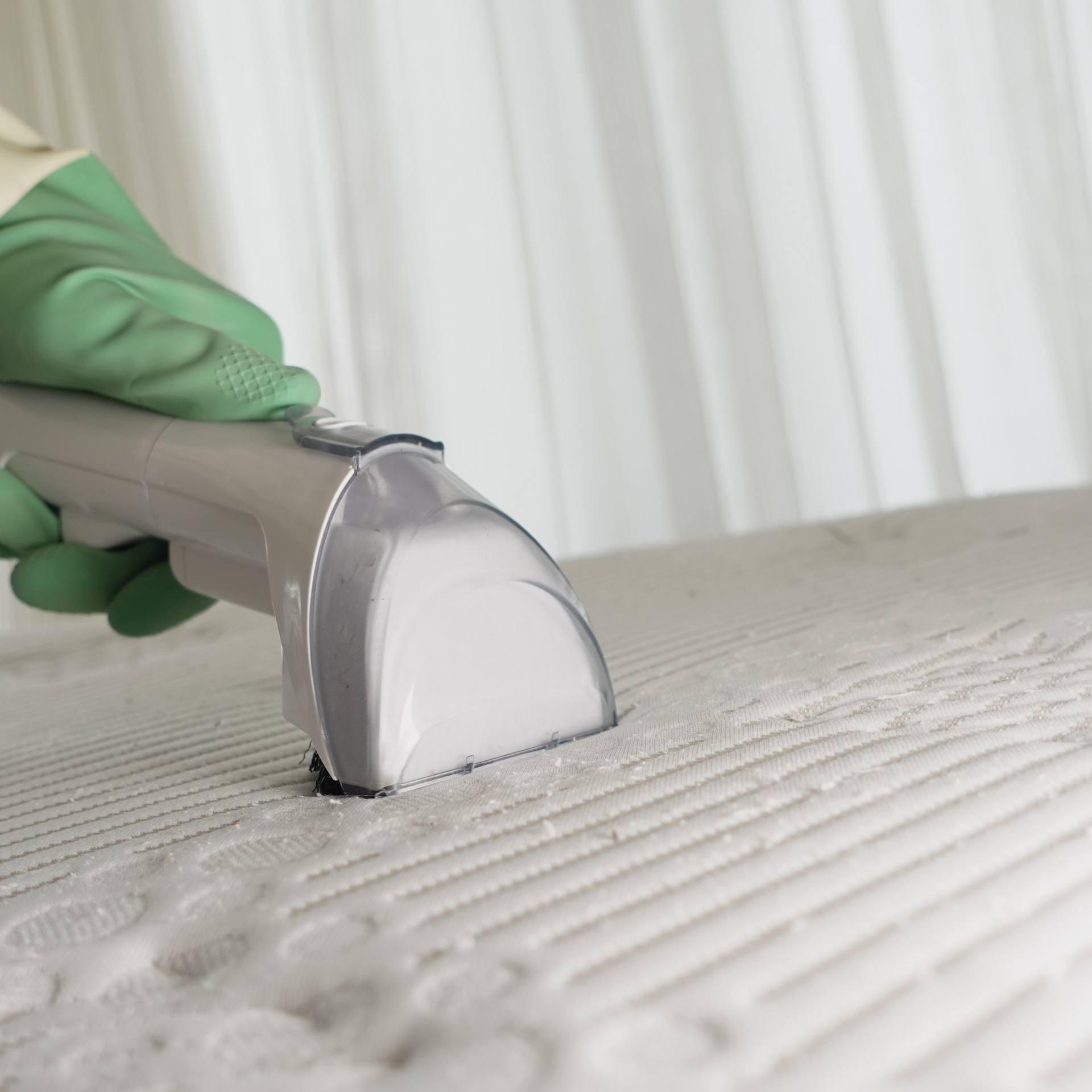Eco Friend Carpet Clean- A person cleaning a white mattress with a handheld vacuum, wearing a green glove.