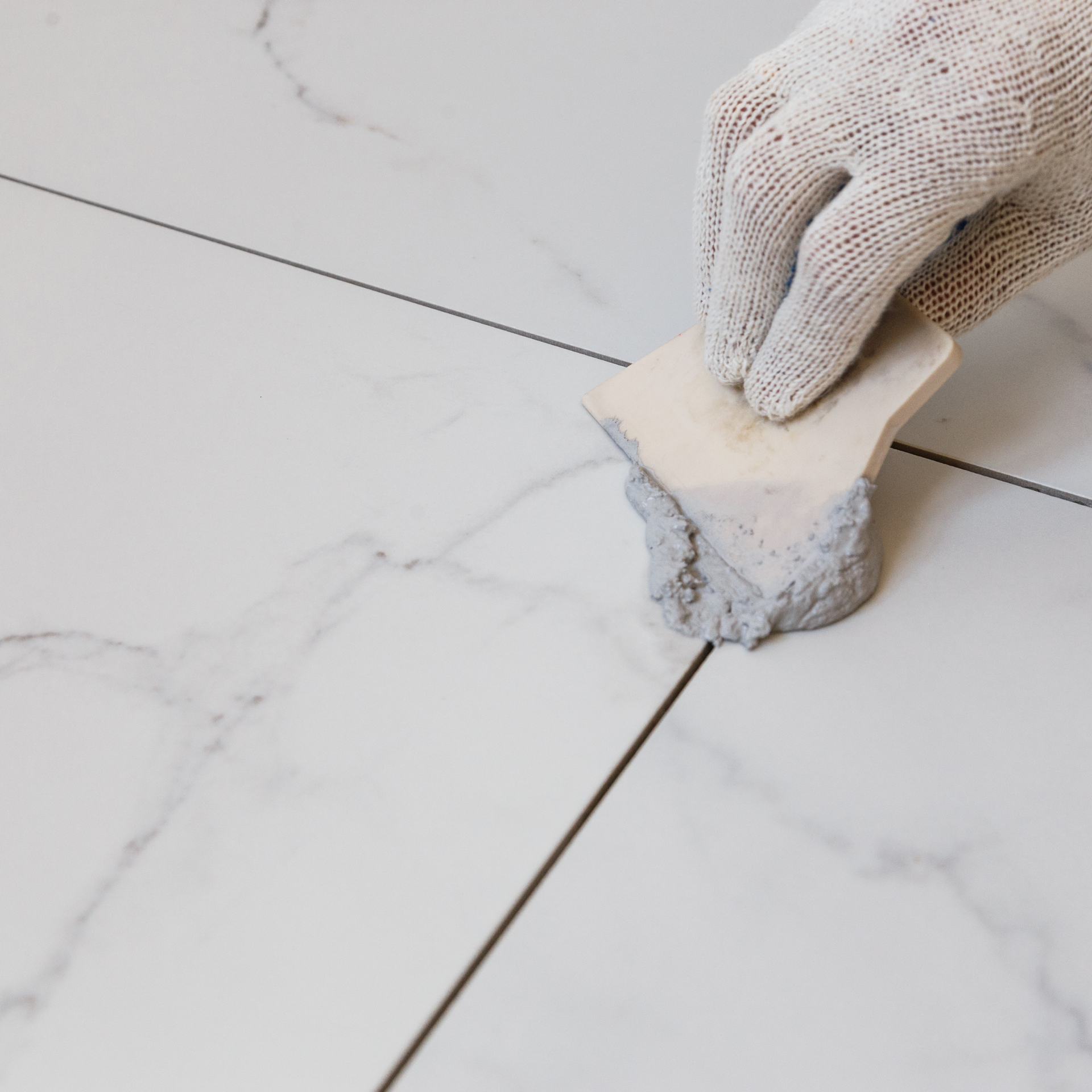 Eco Friend Carpet Clean - Gloved hand using a grout float to smooth gray grout between white marble tiles.