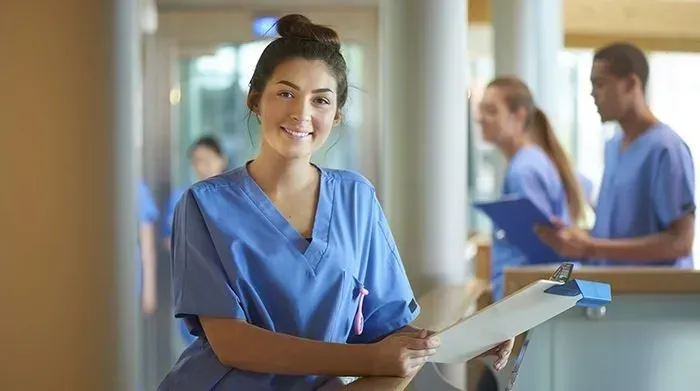 A nurse is smiling while holding a clipboard in a hospital hallway.