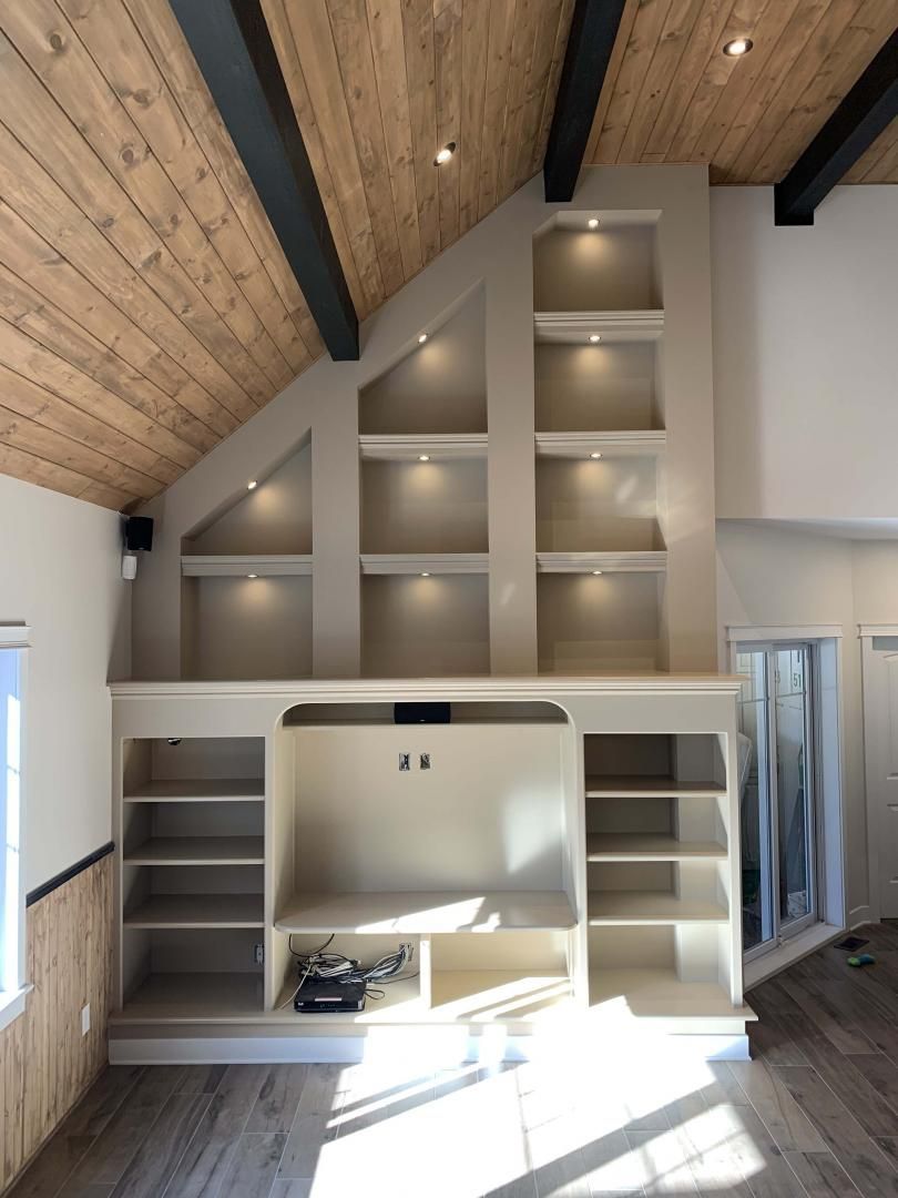 Étagères encastrées et meuble TV sur un mur beige, éclairage encastré, plafond lambrissé et une porte.