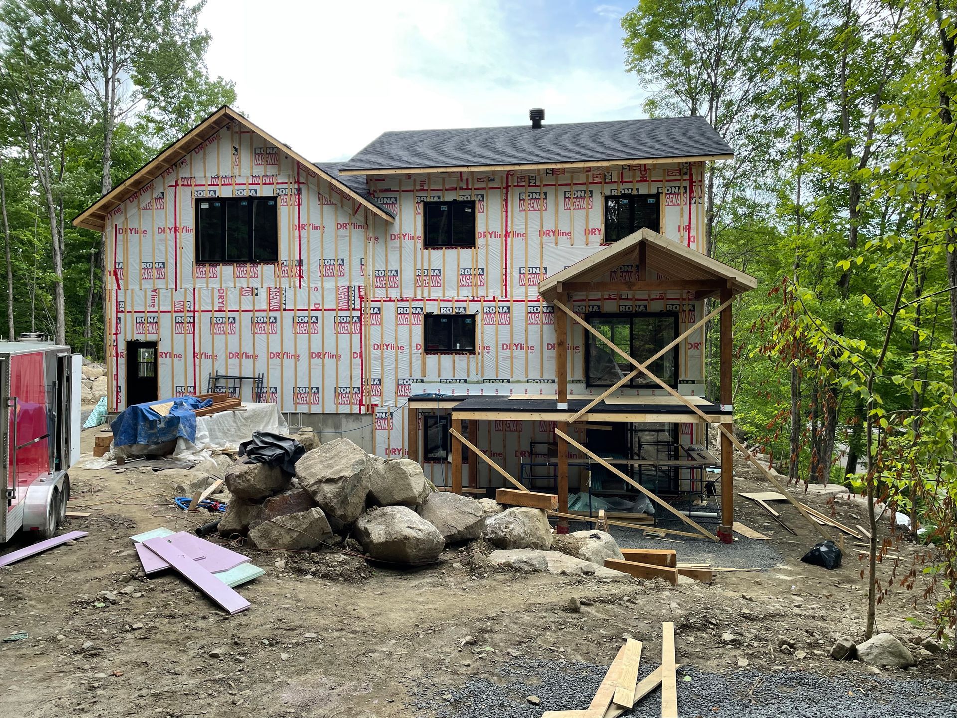 Maison en construction avec charpente apparente, papier goudronné et fondations en pierre. Terrasse et forêt visibles.