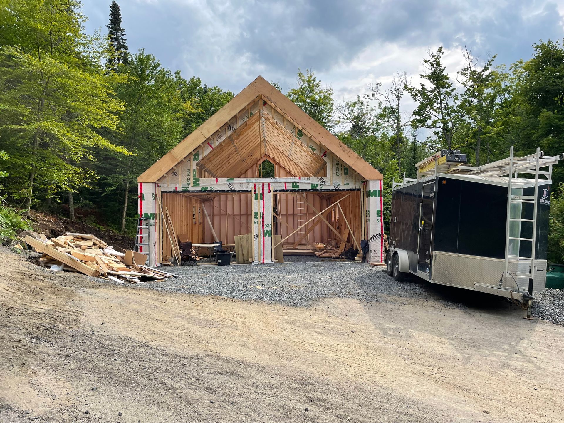 Garage en construction avec ossature bois, deux travées, allée en gravier et une remorque.