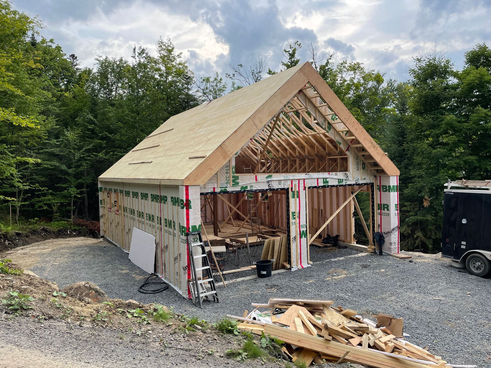 Garage en construction, structure en bois avec toiture partiellement achevée ; allée de gravier, arrière-plan boisé.