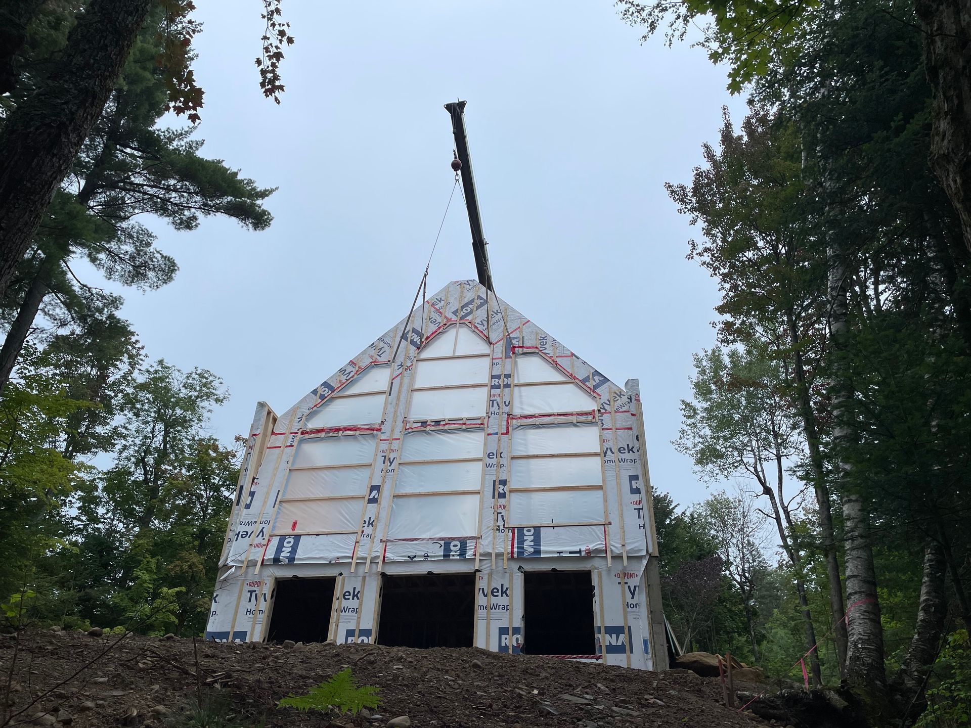 Un bâtiment à ossature blanche, partiellement construit, avec une grue, dans une zone boisée sous un ciel couvert.