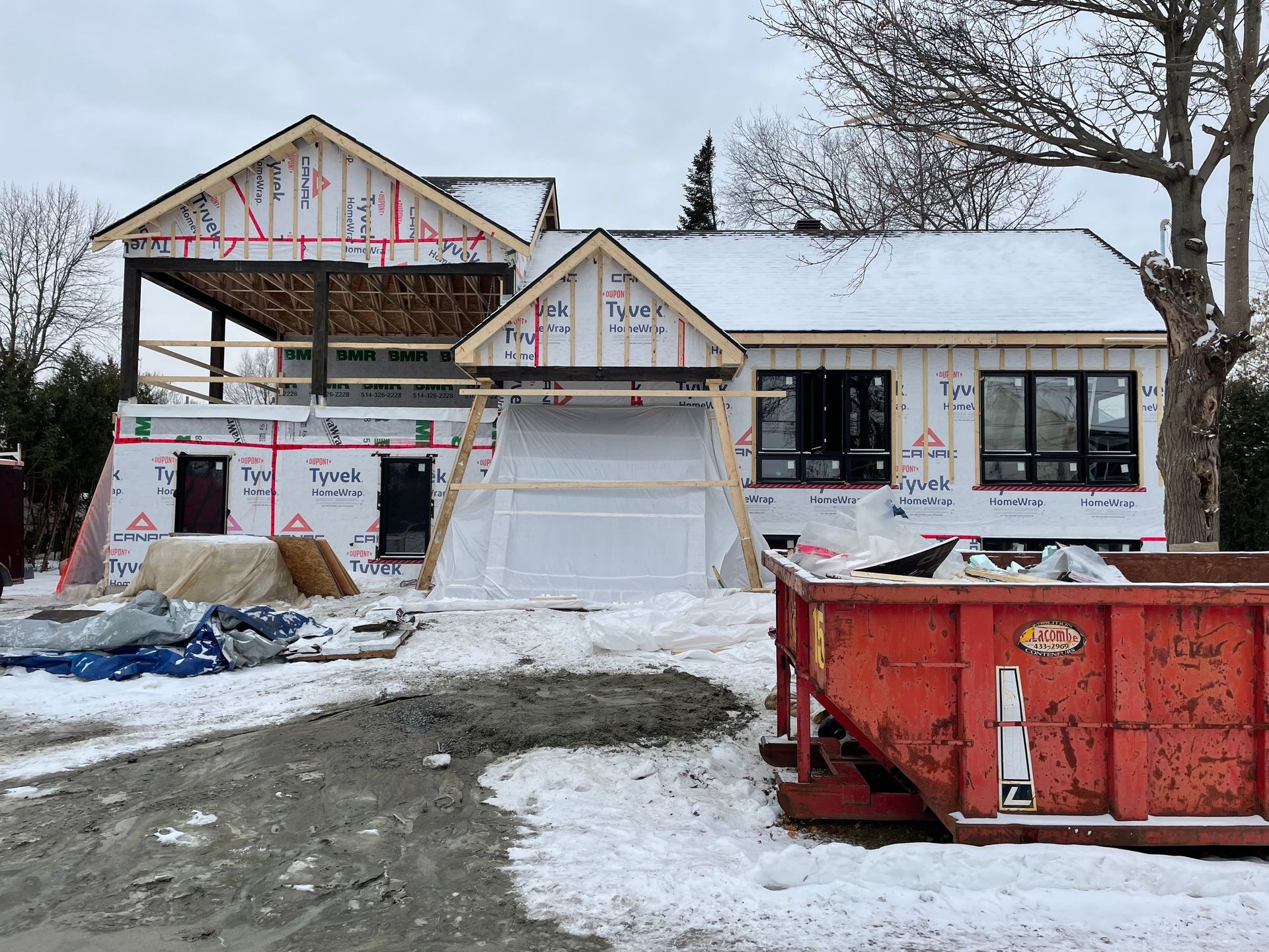 Maison en construction, charpente apparente, recouverte de bâches blanches et de neige. Une benne à ordures rouge se trouve au premier plan.