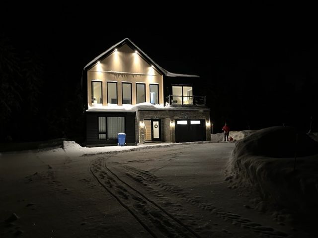Maison à deux étages illuminée la nuit, dans un décor enneigé. Des traces de pas mènent à l'entrée. Une personne se tient à proximité.