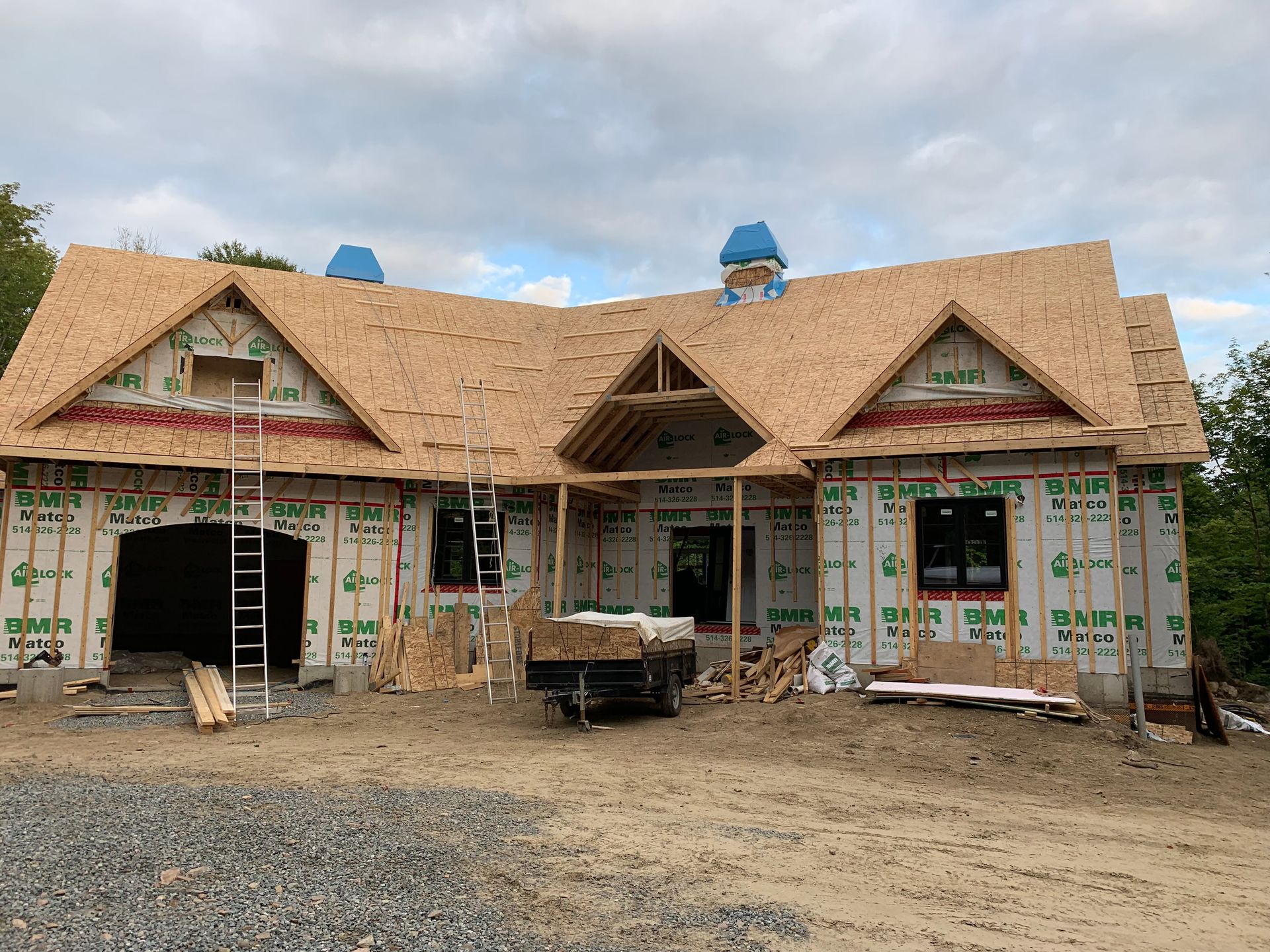 Maison en construction, bardage inachevé, toiture partiellement recouverte de bardeaux, avec échafaudages, située dans un environnement extérieur couvert.