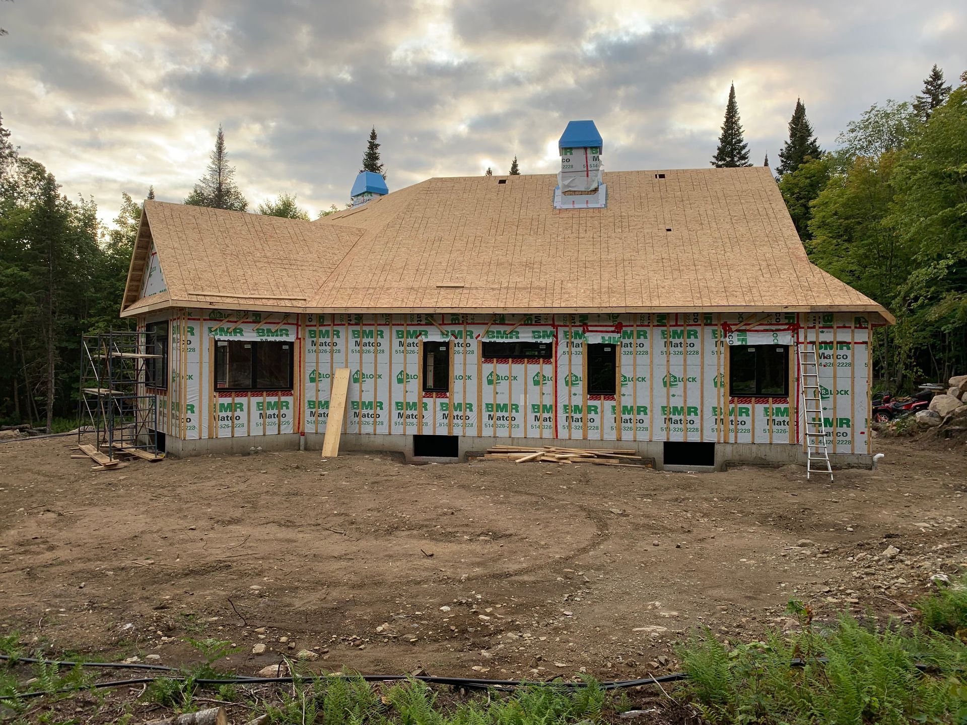 Maison en construction avec toit et murs en bois apparents ; entourée de terre et d'arbres.