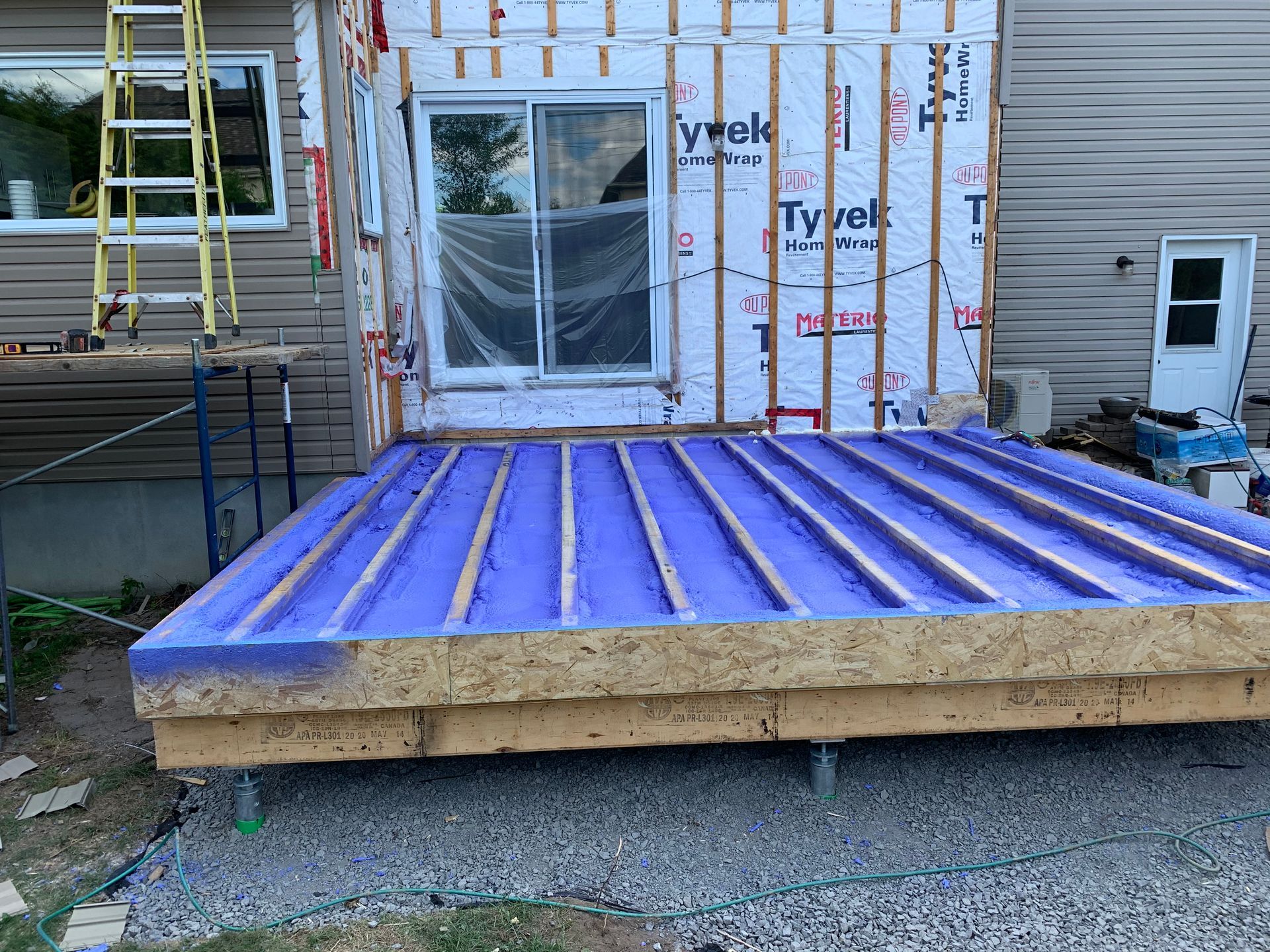 Chantier : Une terrasse en construction, avec une couche imperméable violette sur du bois, près d'une maison avec une porte coulissante en verre.