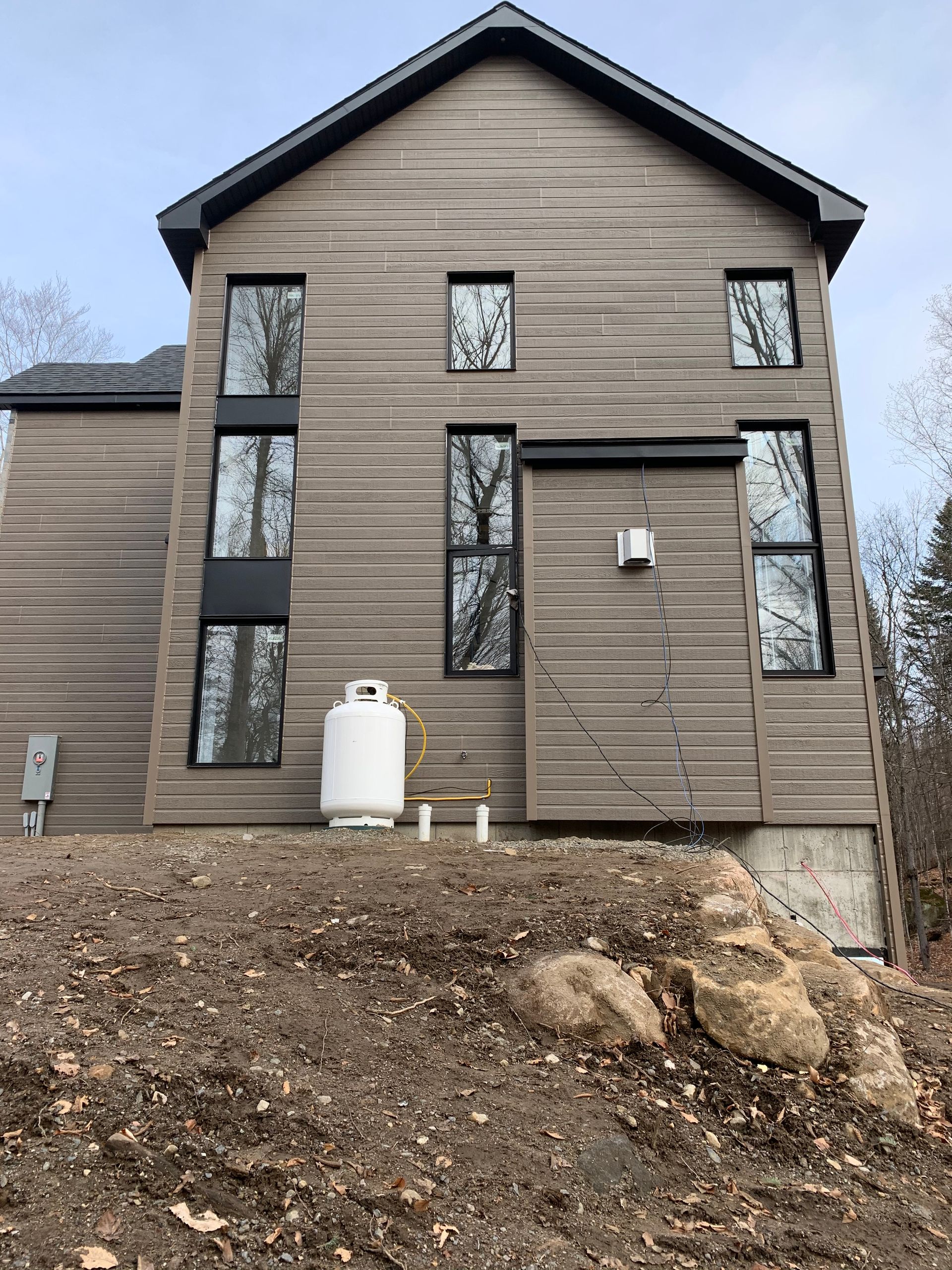 Maison à deux étages avec bardage horizontal gris, fenêtres à cadre noir et réservoir de propane.