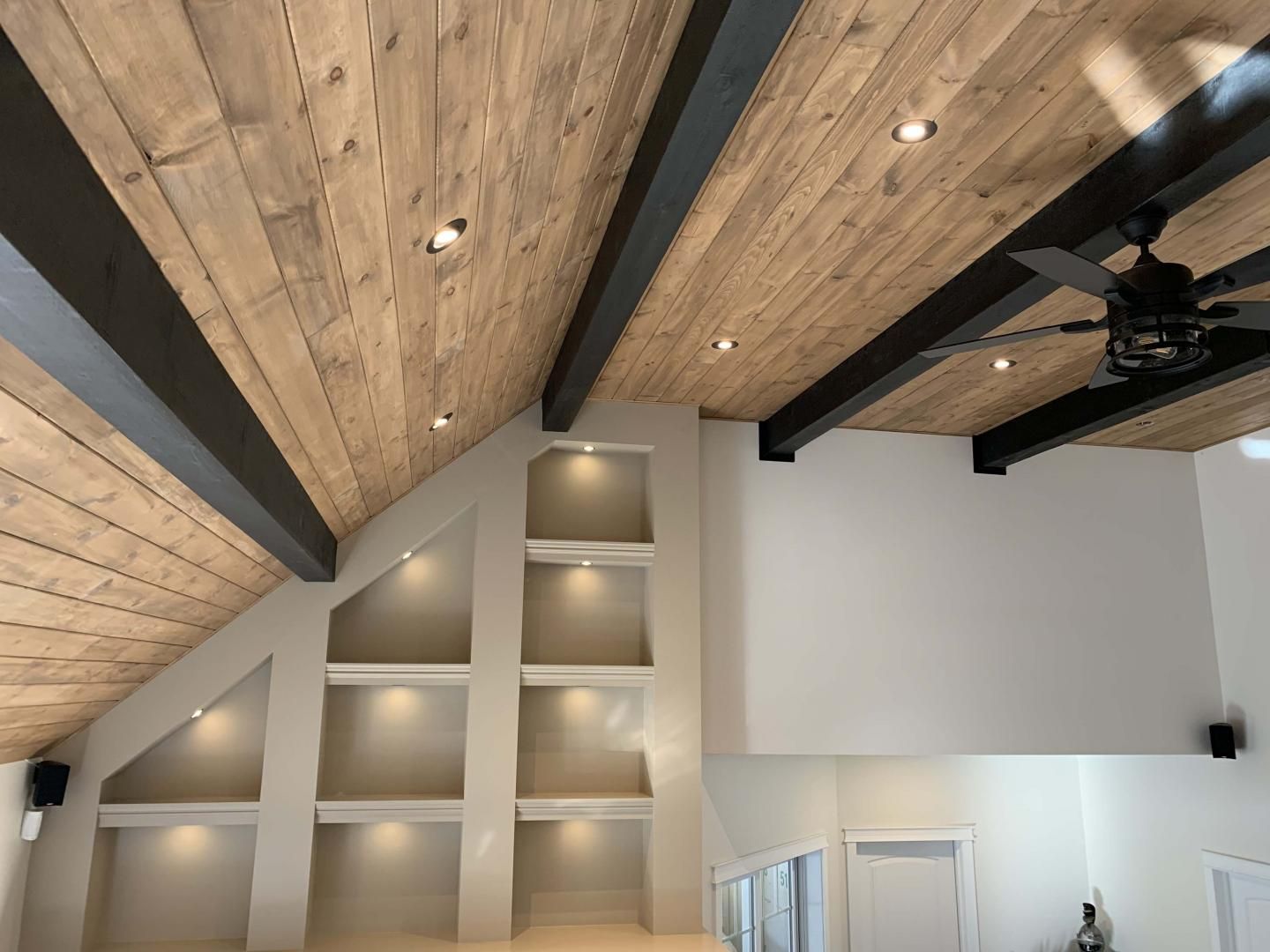 Plafond voûté avec lambris, poutres noires, éclairage encastré et étagères intégrées éclairées par le haut.