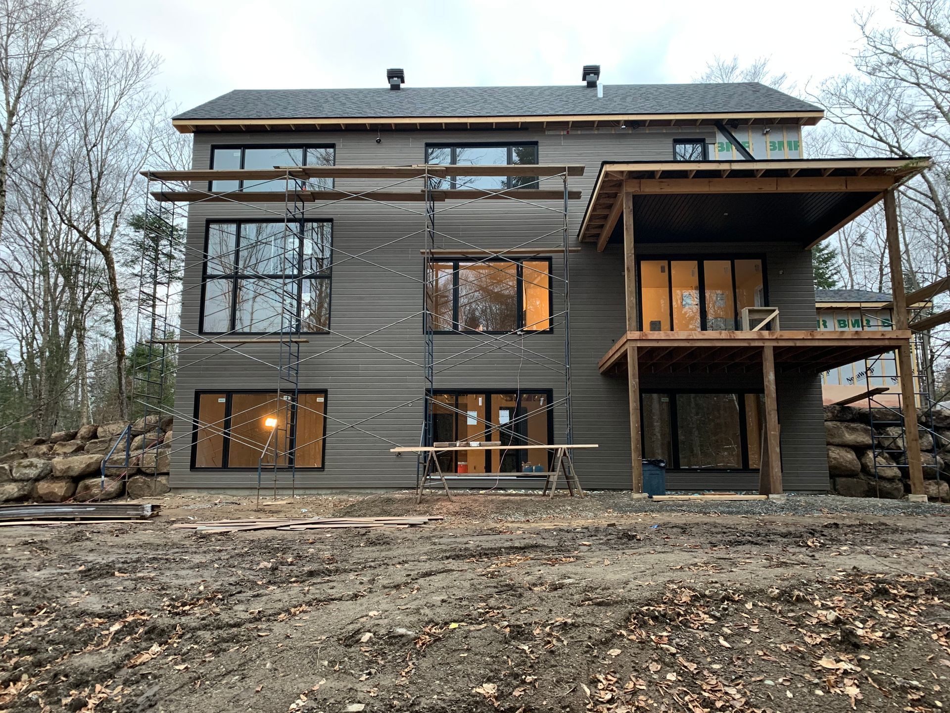 Maison moderne de deux étages avec de grandes fenêtres, un revêtement extérieur gris et une terrasse en bois, située dans une zone boisée.