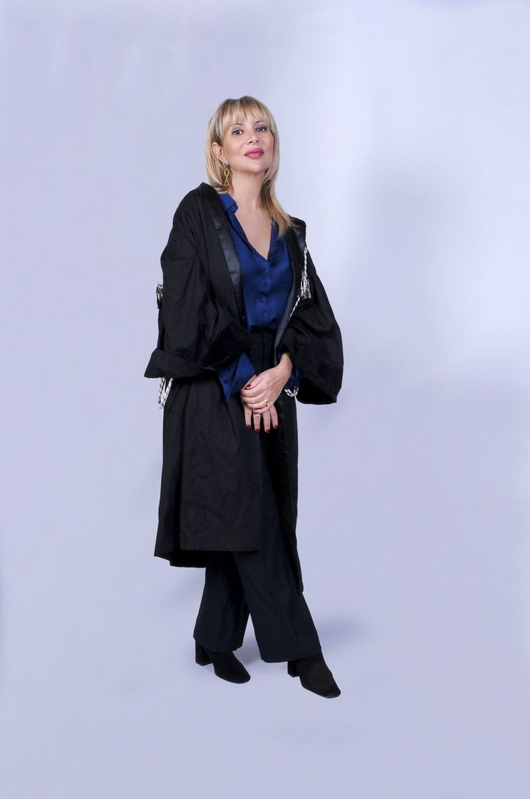 Persona con cappotto nero e maglia blu in piedi davanti a uno sfondo grigio chiaro in studio.