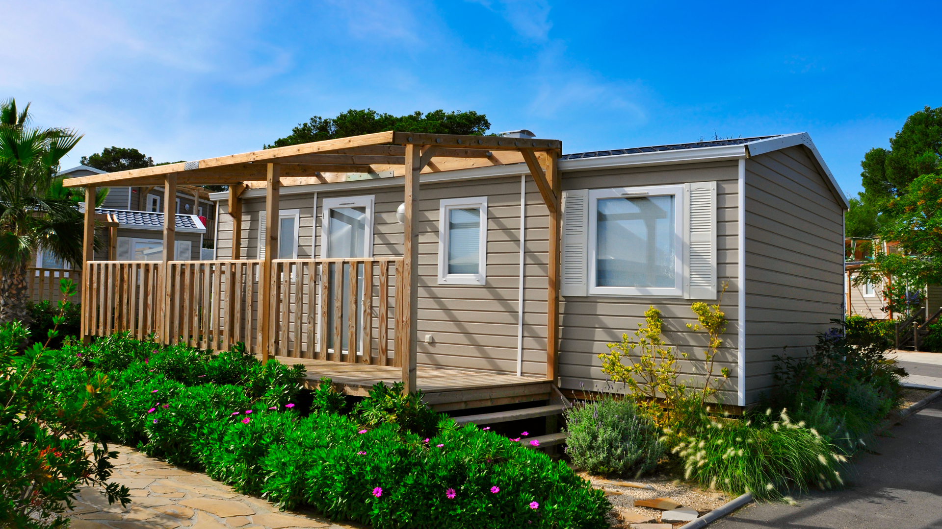 A mobile home with a large deck is surrounded by flowers and bushes.