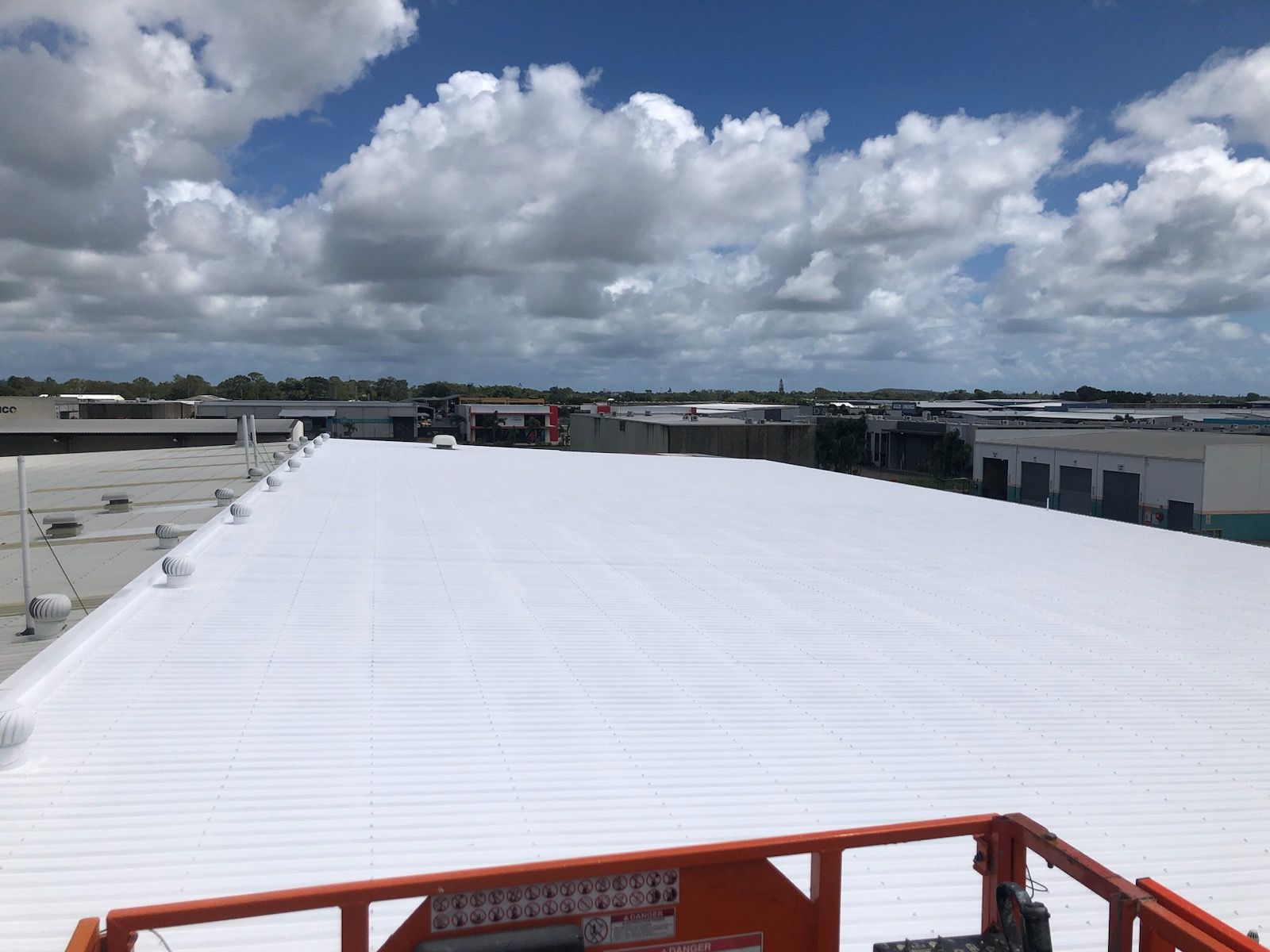 White commercial roof with fluffy clouds overhead. An orange lift is in the foreground. — Depinto Agencies In North Mackay, QLD