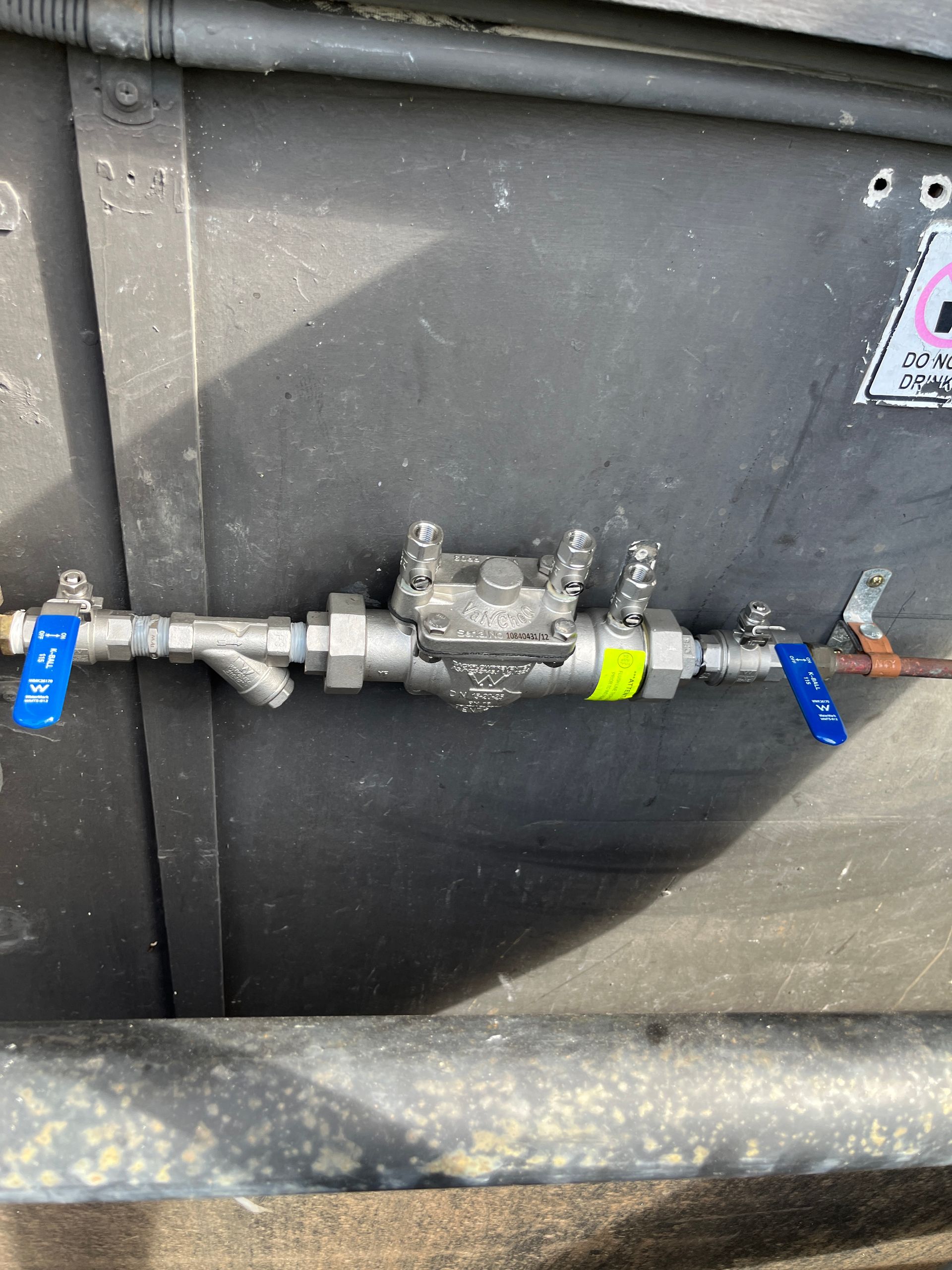 Metal pipe valve assembly with blue-handled shutoff valves mounted on a dark wall outdoors — Depinto Agencies In North Mackay, QLD
