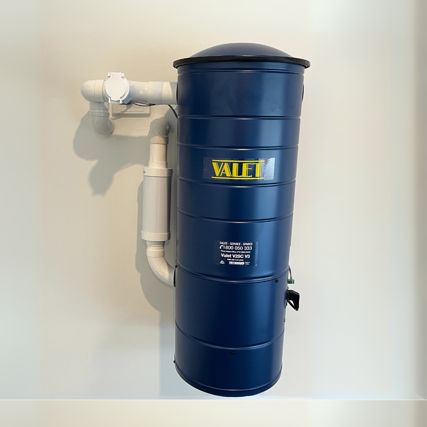 Blue wall-mounted vacuum unit with yellow label and white piping on a beige wall — Depinto Agencies In North Mackay, QLD