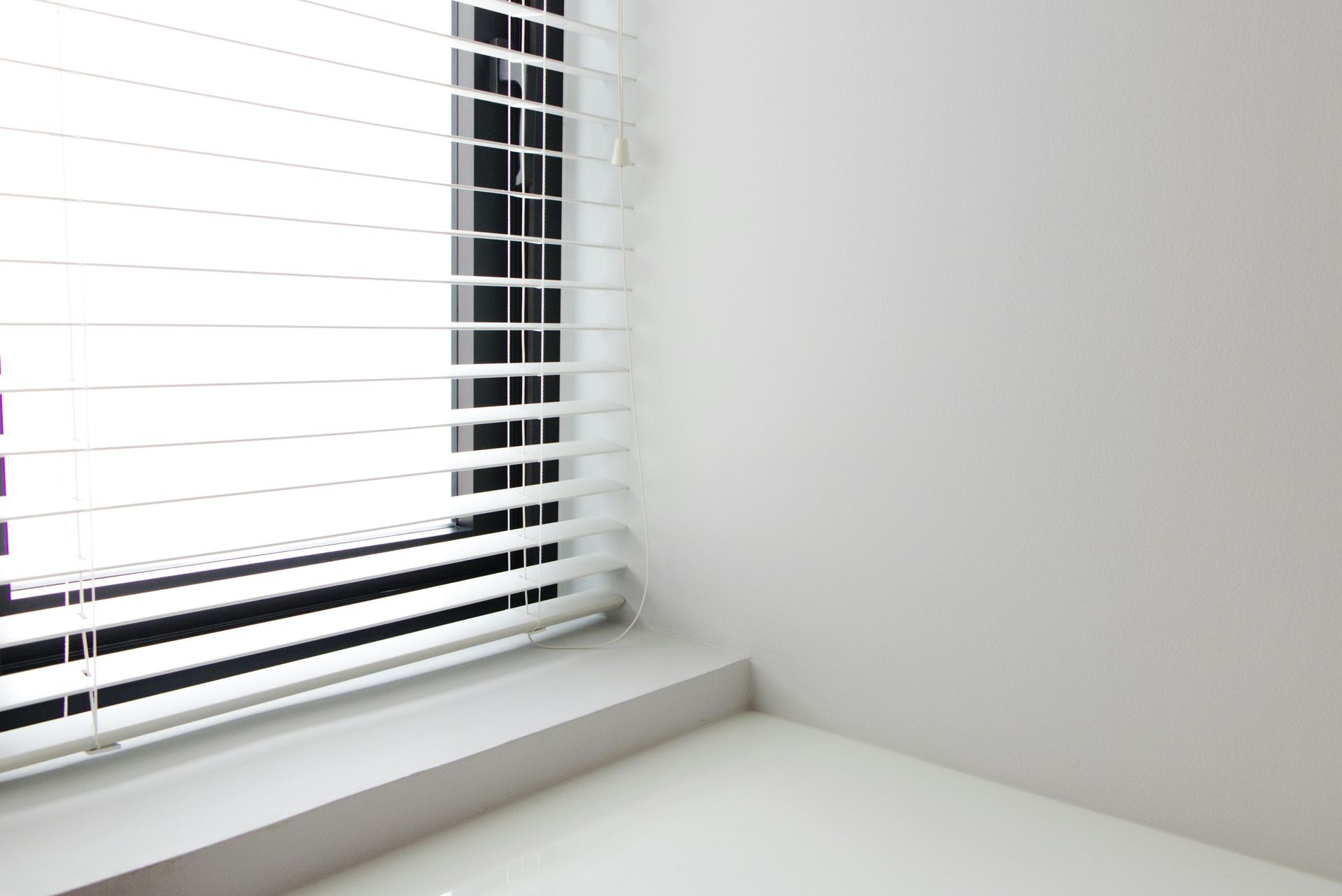 White windowsill beside a window with horizontal blinds, casting light into a plain corner wall.