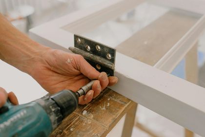 Hands using a drill to fasten a metal hinge on a white wooden frame.
