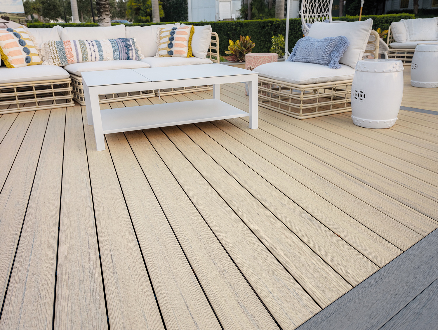 Outdoor lounge area with white wicker furniture, patterned pillows, and a coffee table on a light-colored wooden deck.