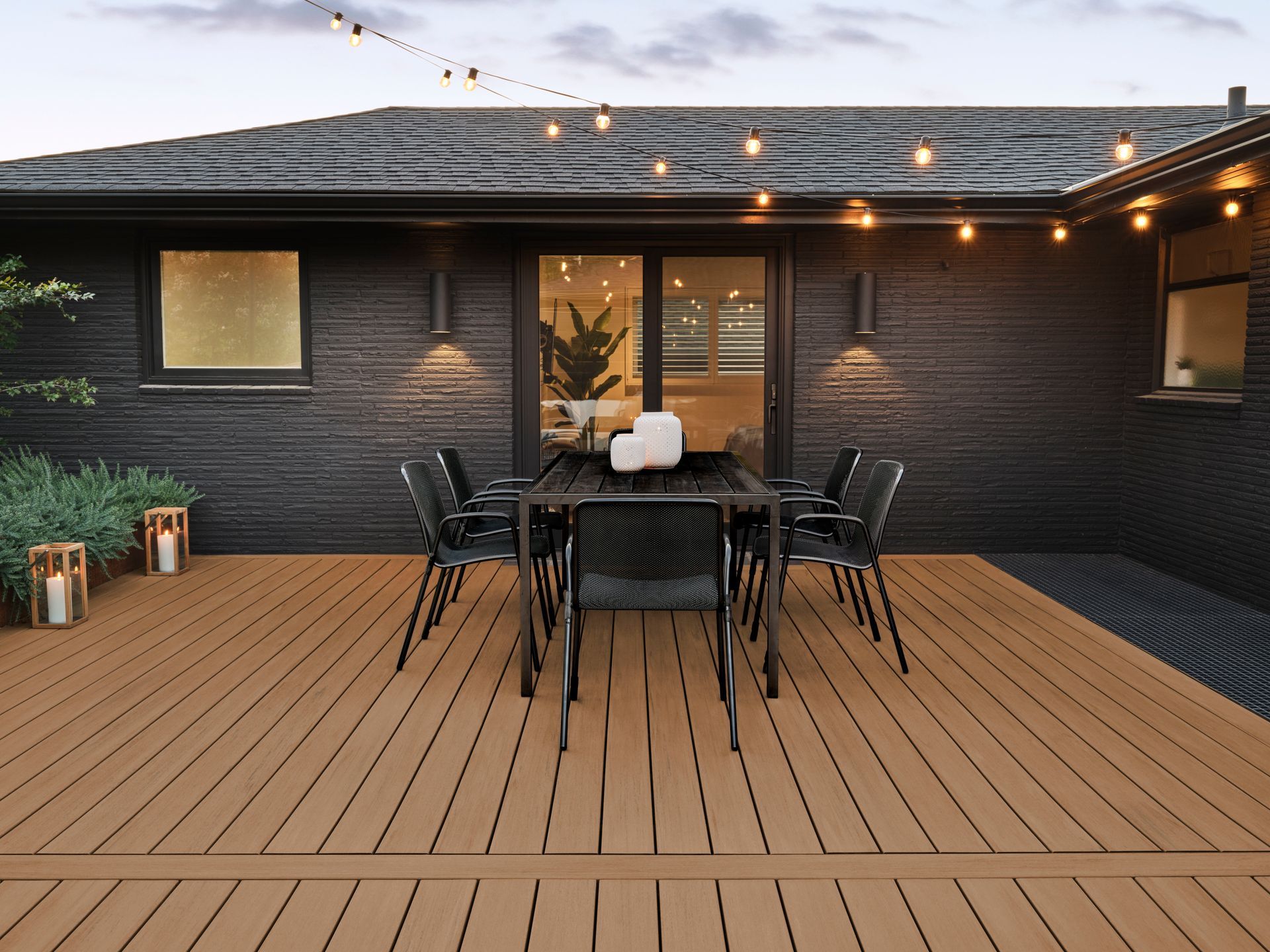 Backyard deck with outdoor dining table and chairs under string lights beside a dark brick house