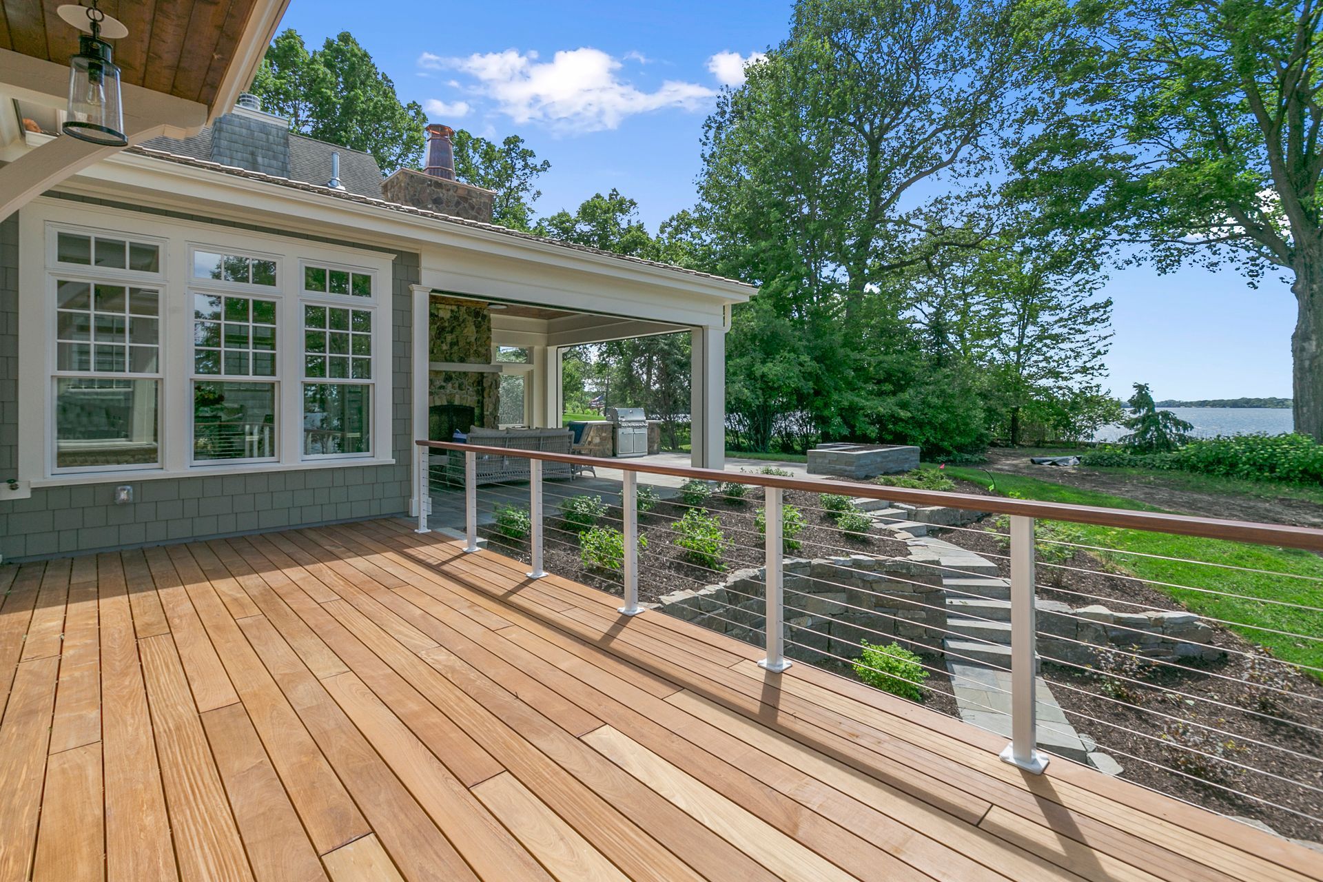 Wooden deck beside a house, with white railing, potted plants, and a view of trees and water beyond.
