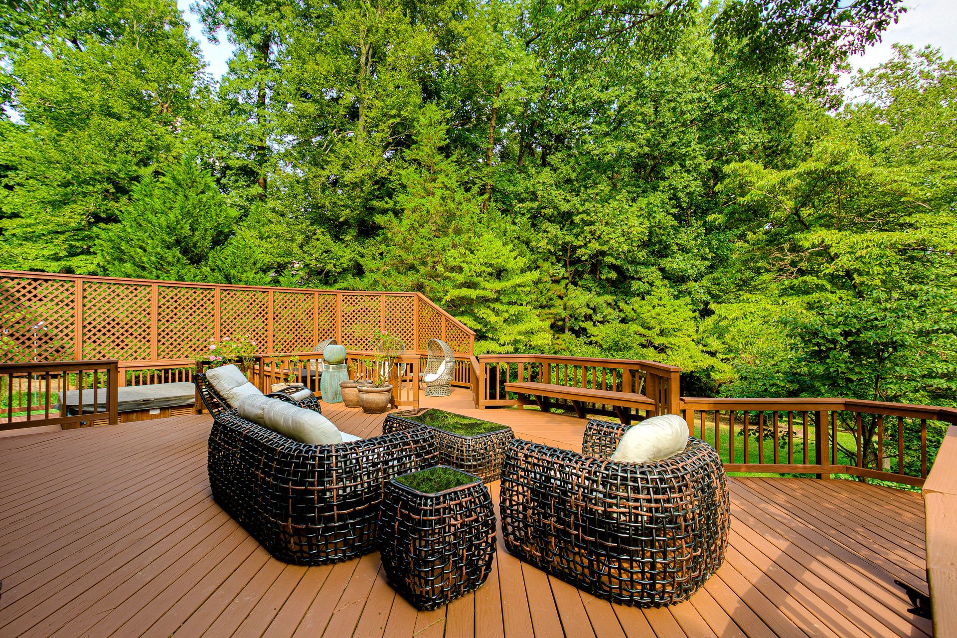Wooden deck with black wicker seating and table, surrounded by lush green trees and a lattice fence.