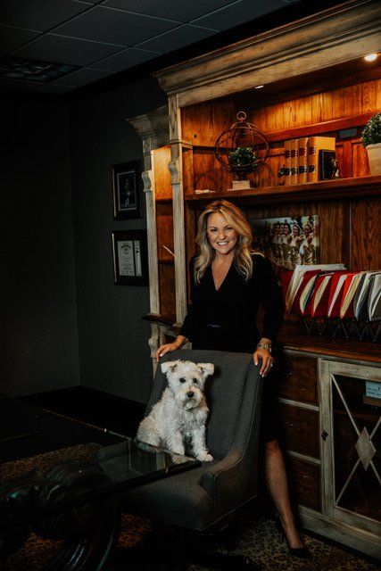 Woman standing beside a gray armchair with a dog in a warmly lit room with wooden shelves.